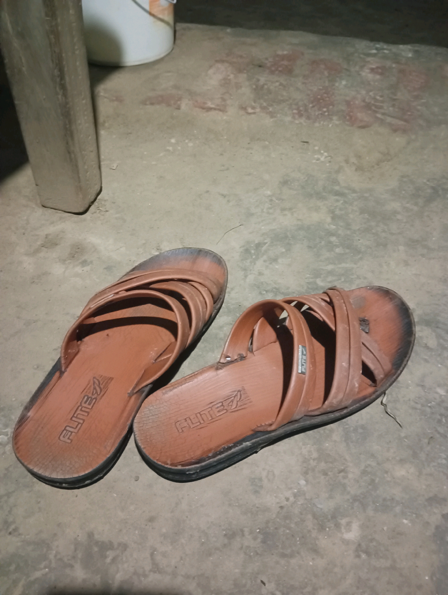 A pair of brown FLITE brand sandals are on a concrete floor. The sandals are worn and have visible scuff marks and dirt. The right sandal is positioned in front of and slightly to the right of the left sandal. In the background, a portion of a white bucket with an orange rim is visible, along with a section of a rough, textured surface that appears to be brickwork. To the left of the sandals is the base of a rectangular, light-colored object that resembles a pillar or support structure. The overall environment suggests an outdoor or unfinished indoor space, possibly in a residential area in Kesari, India, given the context of the provided information.
