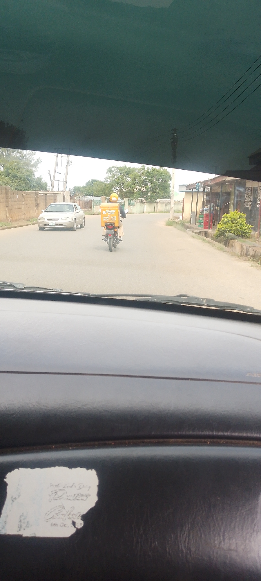 The image captures a street scene in Jos, Nigeria, viewed from inside a vehicle. In the foreground, the dark, textured dashboard of the vehicle is visible, with a crumpled white sticker on the left side featuring handwritten text.
The middle ground shows a paved street with a silver sedan driving away from the viewer on the left side of the road. Further ahead, a person on a red motorcycle is also moving away from the viewer, in the right lane. The motorcycle is equipped with a large yellow cargo box on the back, branded with the word "Speedaf" in black lettering, and additional text below, including what appears to be a phone number. The rider is wearing a yellow helmet.
The background depicts a residential or commercial area with buildings, trees, and utility poles with overhead wires. The overall environment suggests an urban setting with moderate daylight.