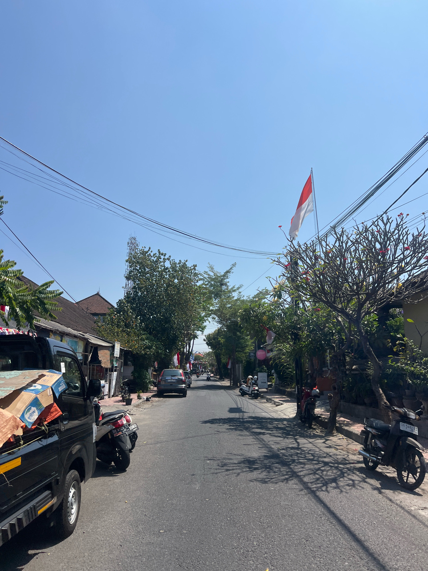 The image shows a street scene in Denpasar, Indonesia, under a clear blue sky. The street is lined with buildings and trees. There are several vehicles present, including a black pickup truck in the foreground loaded with boxes, a silver car in the middle distance, and several motorcycles parked along the side of the road or being ridden. Red and white Indonesian flags are visible, suggesting a national celebration or commemoration. The environment appears to be a typical urban or suburban street with overhead power lines.