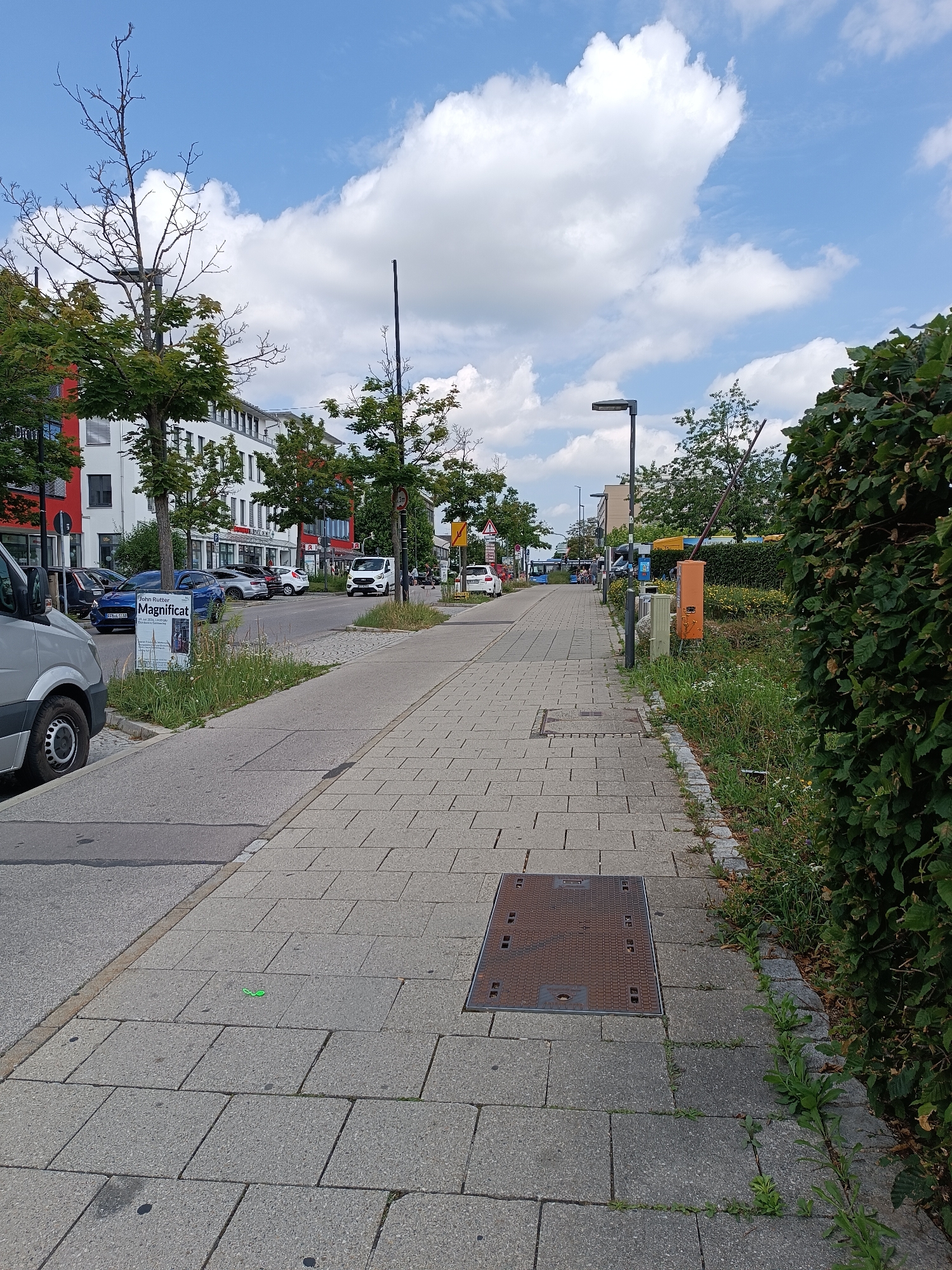The image shows a street scene in Germering, Germany, on a sunny day with scattered clouds. A paved sidewalk runs along the right side of the image, bordered by a green hedge and overgrown vegetation. On the left side of the sidewalk, there is a street with parked cars and a sidewalk, separated by a curb. The street is lined with buildings, some of which appear to be commercial or residential.
In the foreground, on the sidewalk, a manhole cover is visible. Further down the sidewalk, a small green object, possibly a toy or a piece of plastic, is lying on the pavement. A street light is visible on the sidewalk further down. In the background, a white van is driving on the street, and there are other vehicles and people walking.
A sign with the text "John Rutter Magnificat" is posted near the curb on the left side of the image, with some vegetation growing around it. The architecture of the buildings suggests a modern urban environment. The overall scene depicts a typical street in a German town.