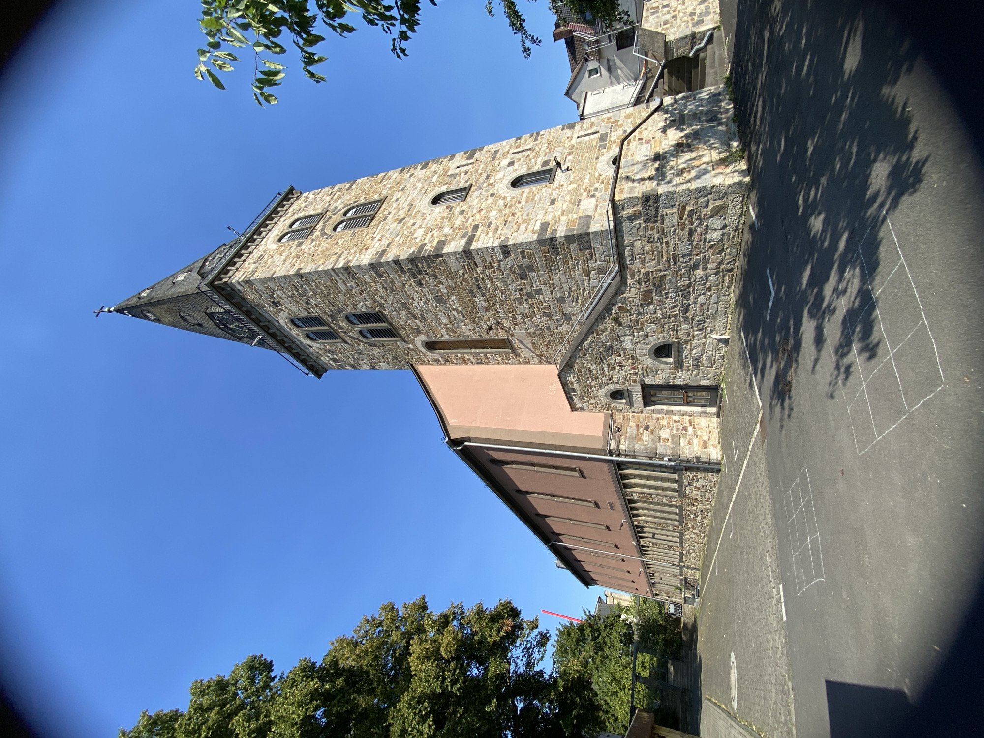 This image shows a church in Altenkirchen, Germany. The church has a tall stone tower with a steeple. The main body of the church is a long, multi-story building with a salmon-pink facade and many windows. The church is surrounded by a paved area with hopscotch squares drawn on the ground. There are trees and other buildings in the background. The sky is clear and blue, indicating a sunny day. The camera angle is low, looking up at the church. There are no people visible in the image.