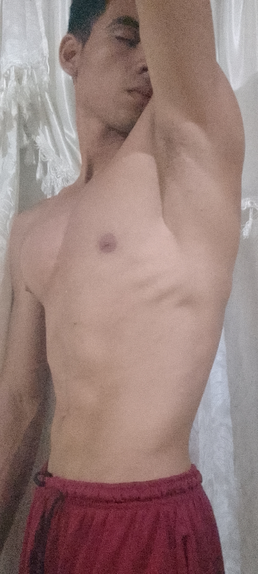 A shirtless man with short, dark hair stands in front of a white curtain. He is wearing maroon shorts. The location is unknown.