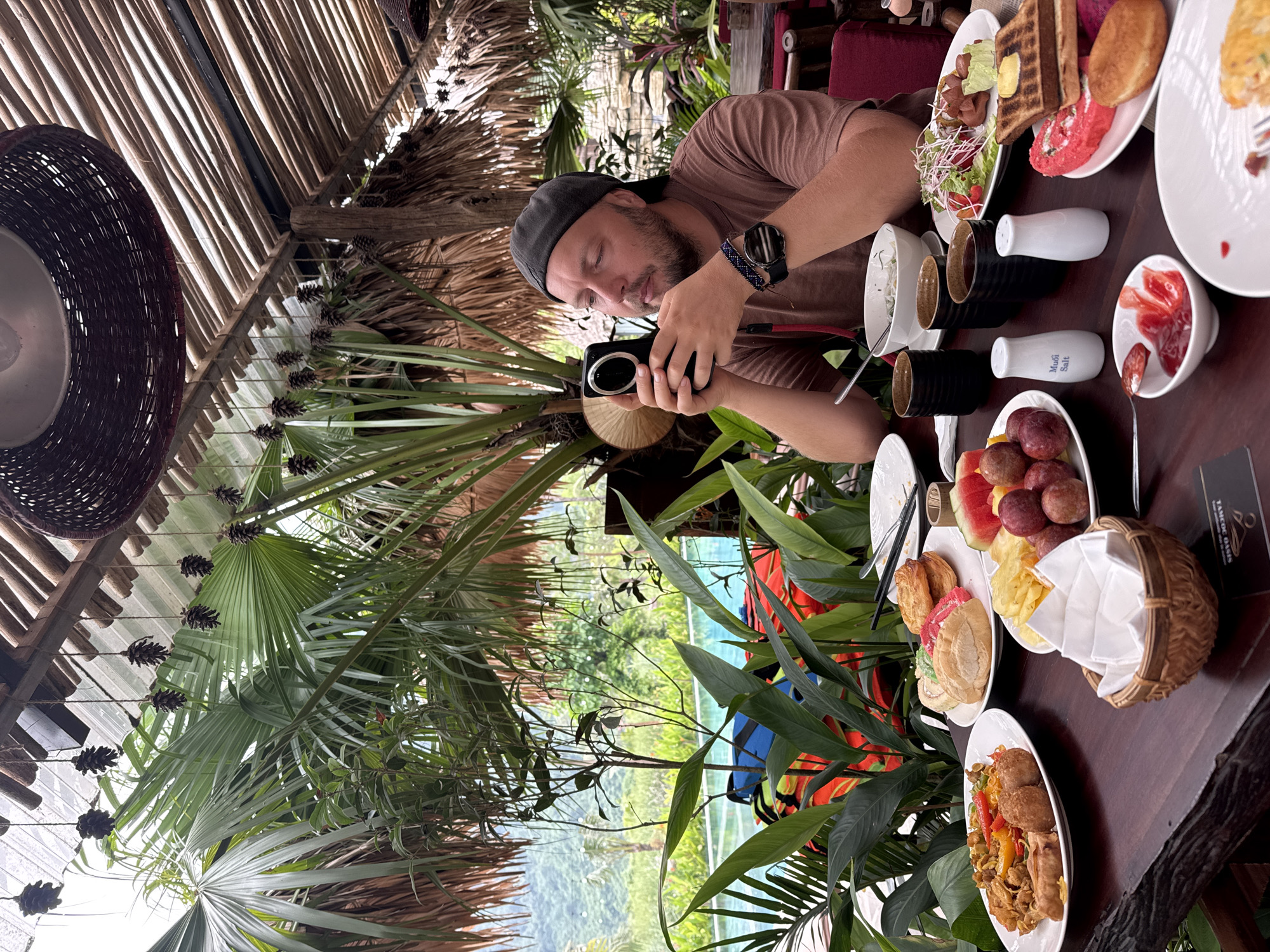 A man wearing a grey baseball cap and a brown t-shirt sits at a wooden table laden with food. He is holding a black camera-like device to his face, appearing to be taking a picture. The table is set outdoors, with a swimming pool and lush greenery visible in the background. The man has a beard and is wearing bracelets on his left wrist.
The table features several plates of food, including what appears to be a breakfast spread with pastries, fruit, potato chips, and a salad. There are also small cups, salt and pepper shakers, and a woven basket with napkins. The setting suggests a resort or restaurant with an open-air dining area, characterized by a thatched roof supported by wooden beams and palm fronds. The environment is tropical, with abundant vegetation and a mountain range visible in the distance.
The location is identified as Nam Hoa Lư Ward, Vietnam. The overall scene depicts a man enjoying a meal in a scenic, tropical environment, engaged in the act of photography.