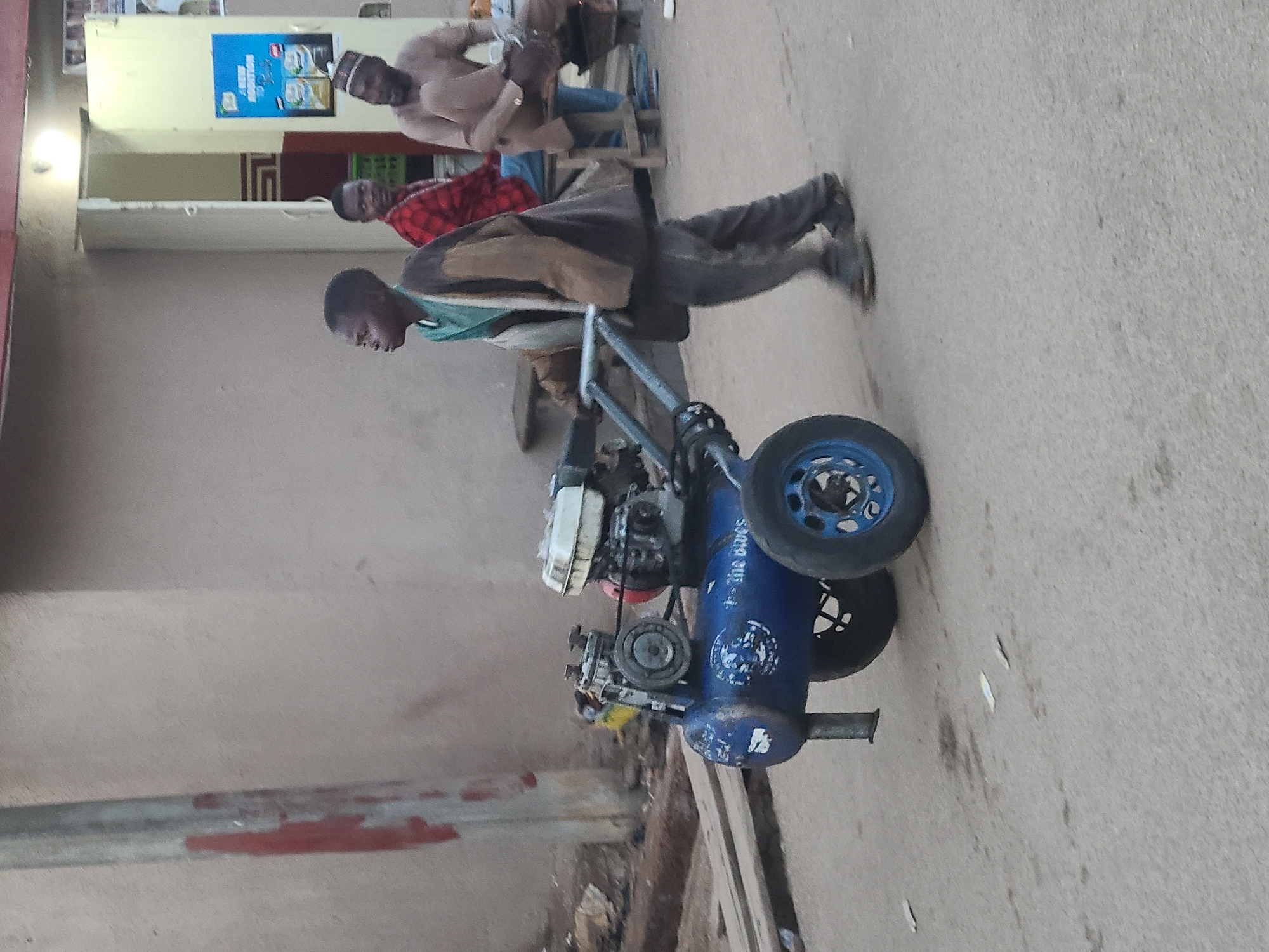 This image captures a daytime street or market scene featuring several men and a prominent air compressor. In the foreground, a young man, dressed in a green shirt, brown jacket, and dark pants, stands alongside a blue portable air compressor. The compressor has an engine unit, two main wheels (one blue, one black), and "AIR COMPRESSOR," "SAWES," and "300-L" visible on its side, along with the partially visible text "the owes." He is looking towards the left of the frame.
Behind him, another man is seated, wearing a red and black checkered shirt. Further in the background, a third man stands near a building, dressed in a light-colored traditional garment and a skullcap. A blue poster is affixed to a wall in the background, displaying the words "NEW BOOSTION" and an image of a product. The ground is paved, suggesting an urban or semi-urban setting. The lighting is soft and diffused, indicating either early morning or late afternoon. The general atmosphere is calm, with people engaged in their daily routines. The attire of some individuals and the general environment hint at a location in a non-Western country.