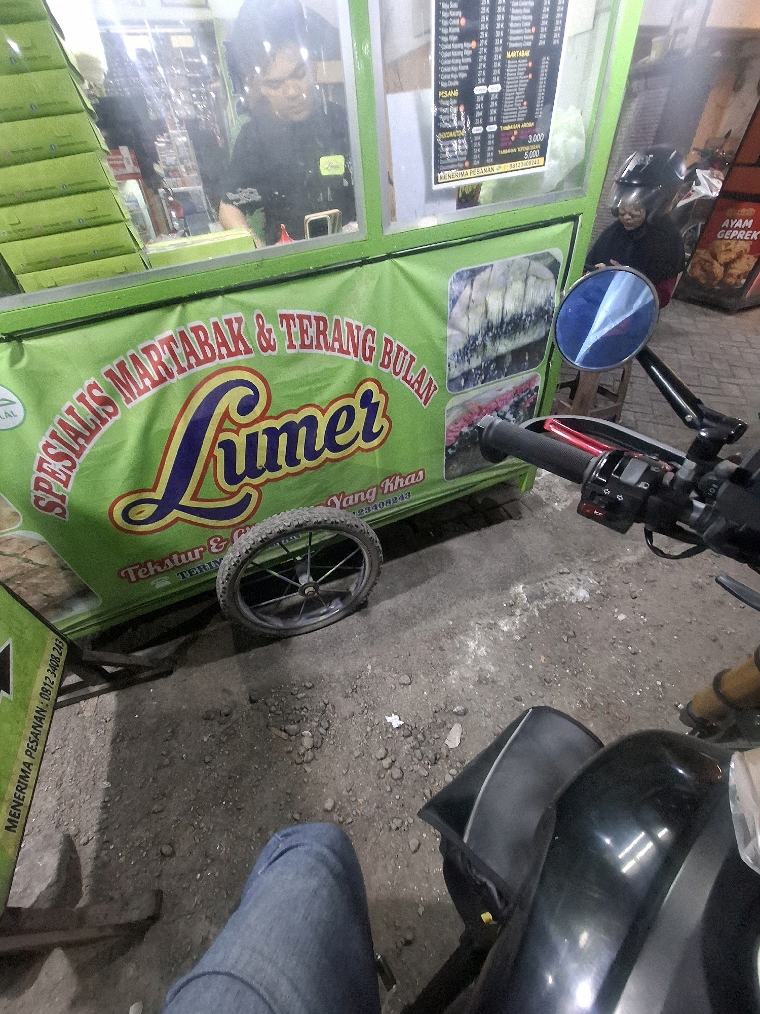 The image shows a street view in front of a food stall in Perumahan Puri Indah, Indonesia. The food stall, which appears to sell martabak and terang bulan, has a green facade with text and images. A person wearing a black shirt and glasses is visible inside the stall, possibly preparing food. A motorcycle is parked in front of the stall, with its handlebars and part of its body visible. A rider wearing a helmet is seated on the motorcycle. The ground is paved with asphalt and pebbles, and there are some scattered debris. In the background, a menu board is displayed, listing various food items and their prices. There is also a sign indicating "AYAM GEPREK" which translates to "crushed chicken," a popular Indonesian dish. The overall scene depicts a typical street food vending environment in Indonesia.