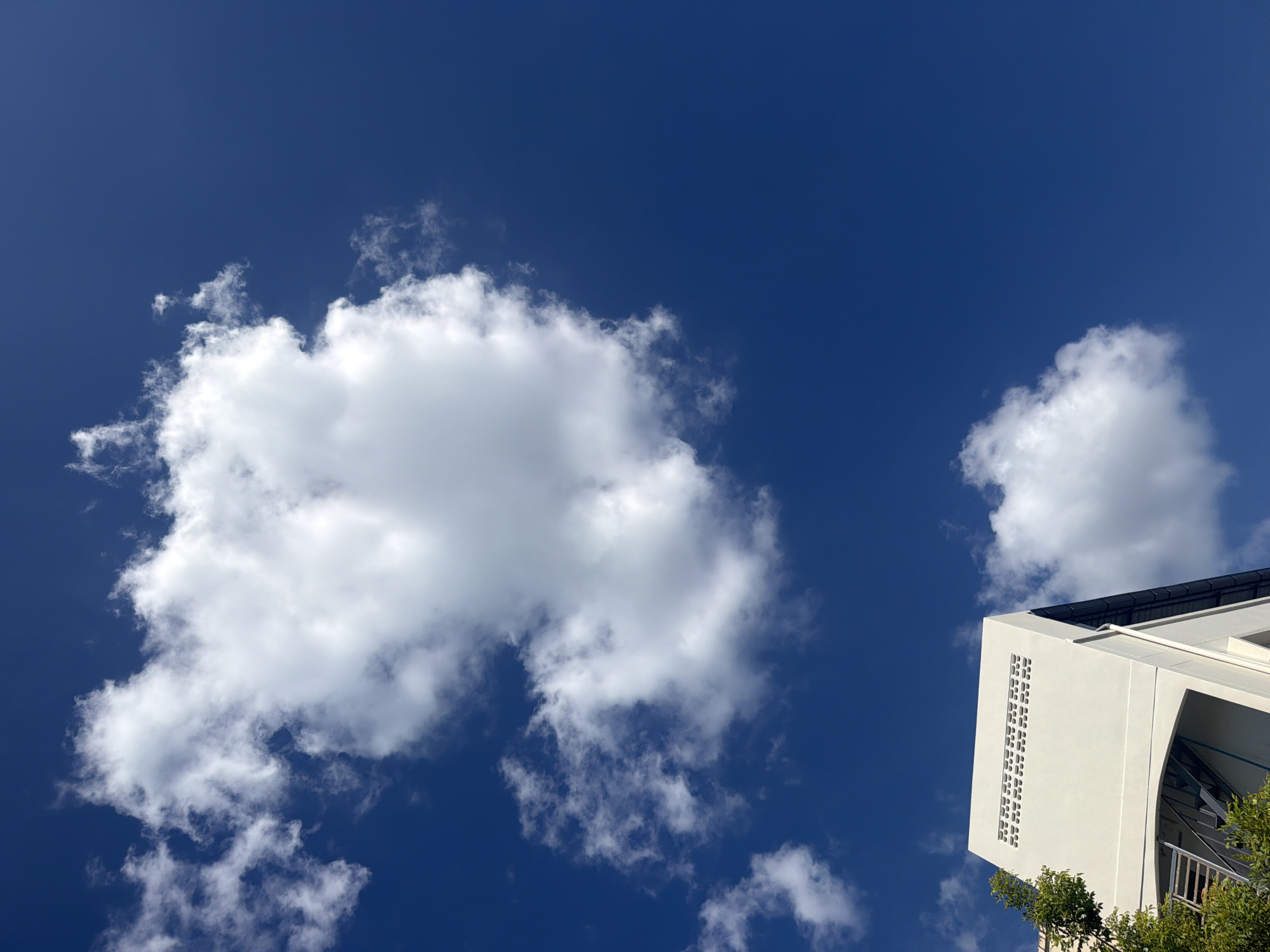 A low-angle, vertical shot captures a bright blue sky with scattered white cumulus clouds. In the lower left quadrant, the upper section of a light-colored building is visible, featuring a row of rectangular vents near its peak and an external metal staircase on its left side. Lush green foliage partially obscures the building from the left. A second, smaller white cloud floats in the sky to the right of the building. The scene is consistent with a daytime view in Baan Chaweng, Thailand.