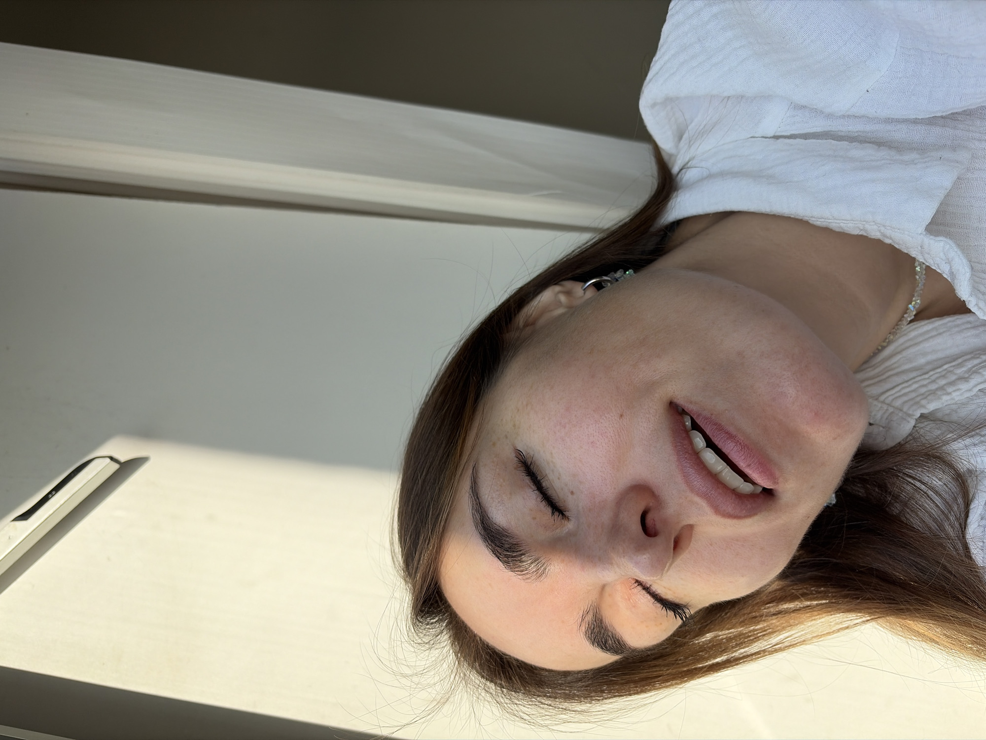 A fair-skinned woman with brown hair and closed eyes is positioned in the lower right portion of the image. She is wearing a white blouse and a crystal necklace. A pen is visible on a white surface in the lower left corner. A light-colored wall with a dark trim is in the background. The location is not discernible from the given image.