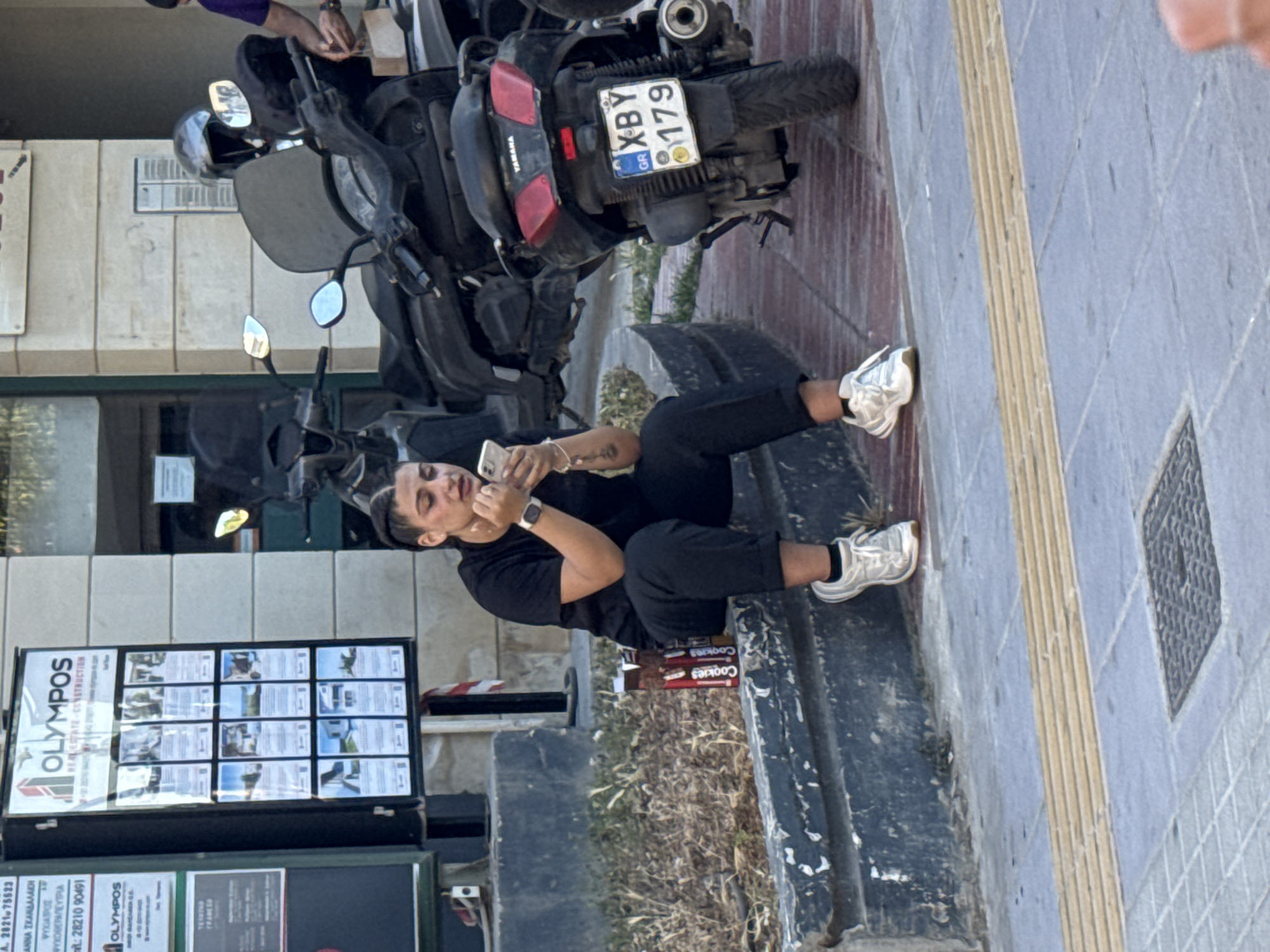 The image depicts a street scene in Chania, Greece. In the foreground, a young woman is sitting on a low stone wall, looking at her phone. She has dark hair styled in braids and is wearing a black t-shirt and black pants, with white sneakers. Two red boxes of "Cookies" are placed on the wall next to her. Behind her, several motorcycles are parked. A blue and white license plate on one of the motorcycles reads "XBY 179 GR". A real estate advertisement board with photos of properties is visible on the left side of the frame, bearing the "OLYMPOS REAL ESTATE - CONSTRUCTION" logo. The ground is paved with light-colored tiles and there is a tactile paving strip indicating a pedestrian walkway. The background shows a building facade and a hint of greenery.