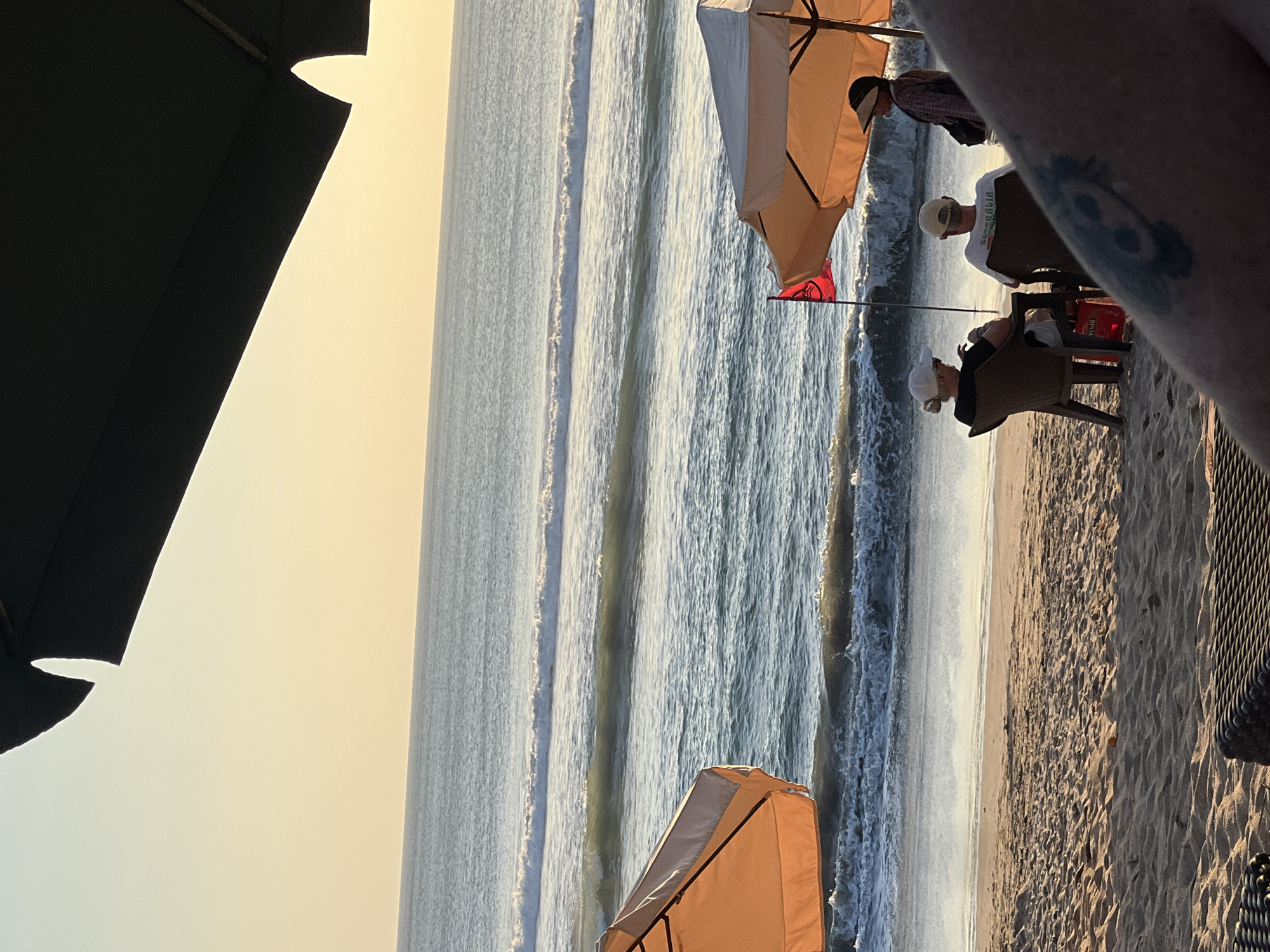 The image shows a beach scene in Kuta, Indonesia. In the foreground, a person's arm with a tattoo is visible, partially obscuring the view of a woven lounge chair. Further back, two people are seated on chairs under an orange umbrella, facing the ocean. A third person stands near the umbrella. A red flag with a symbol is planted in the sand. The ocean has moderate waves, and the sky is a soft yellow from the sunset. The beach is sandy, with footprints and some scattered debris. The scene suggests a relaxed evening by the sea.