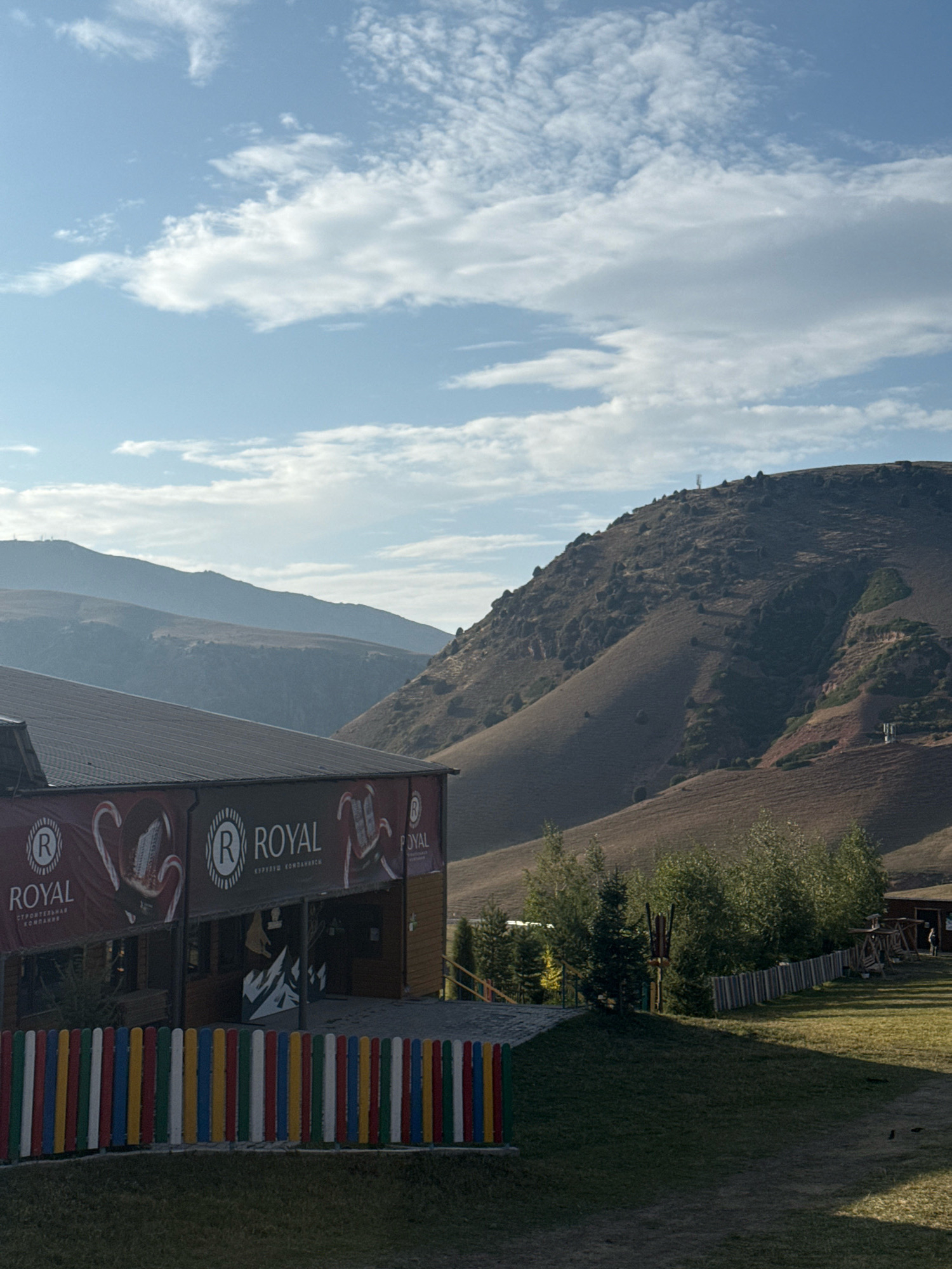 The image depicts a scenic mountain landscape in Chunkurchak, Kyrgyzstan, on a clear day with scattered clouds. In the foreground, a rustic building with a dark-tiled roof is partially visible, featuring a banner with the logo "ROYAL" and what appears to be images of candies. A colorful, multi-striped wooden fence runs along the front of the property. Beyond the building, rolling, brown-toned hills dominate the middle ground, with sparse vegetation and some trees dotting their slopes. In the distance, layers of hazy mountains recede into the background, under a bright blue sky. The overall impression is one of a rural or recreational area situated within a mountainous terrain.