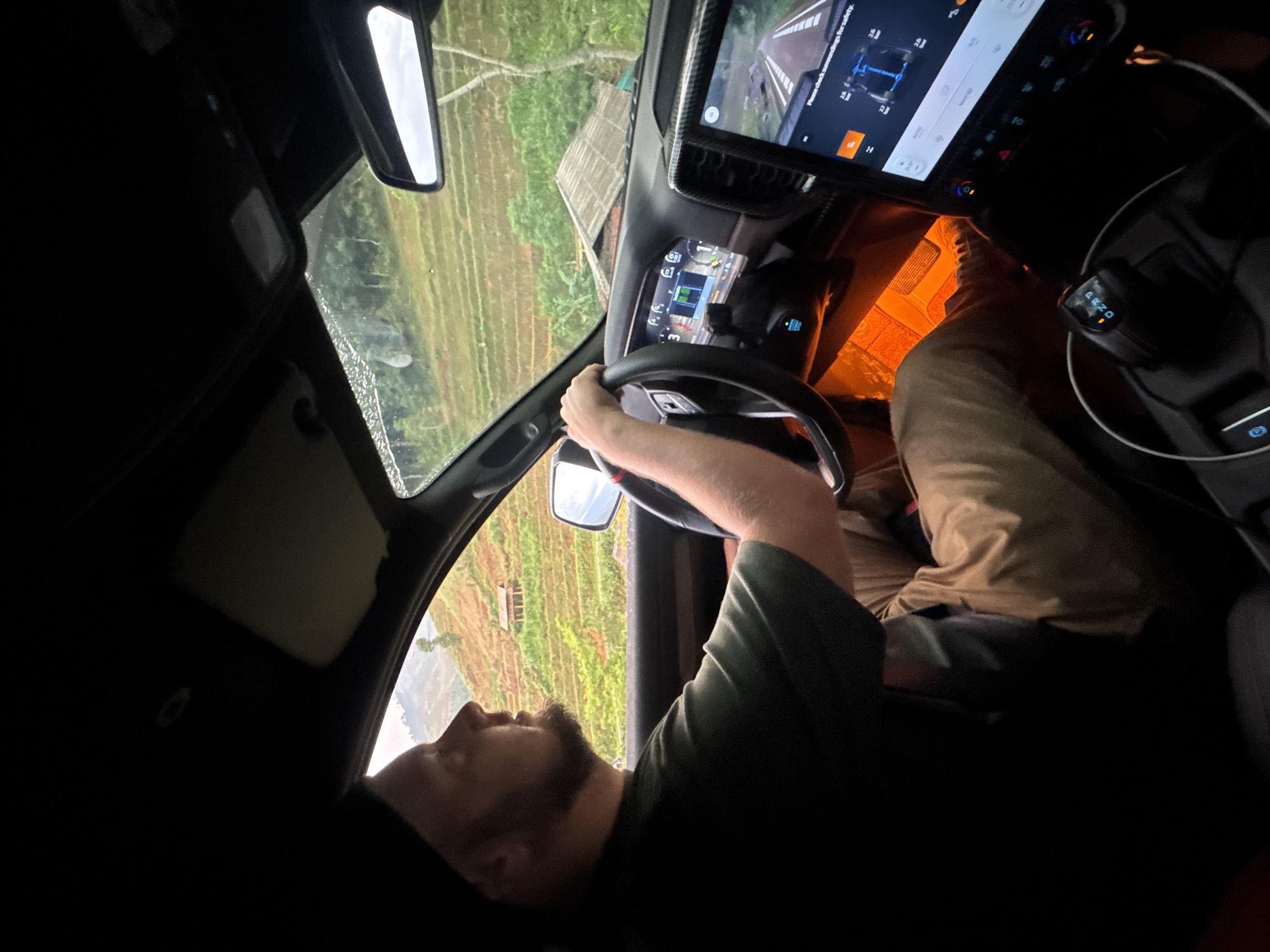 A man is shown driving a car on a road in Dế Xu Phình, Vietnam. The man has a beard and is wearing a dark green shirt and tan pants. The car's dashboard and steering wheel are visible, as is the digital display. The scenery outside the car window features rice paddies, characteristic of the region. The view is from inside the car, and the photo is oriented at an angle.