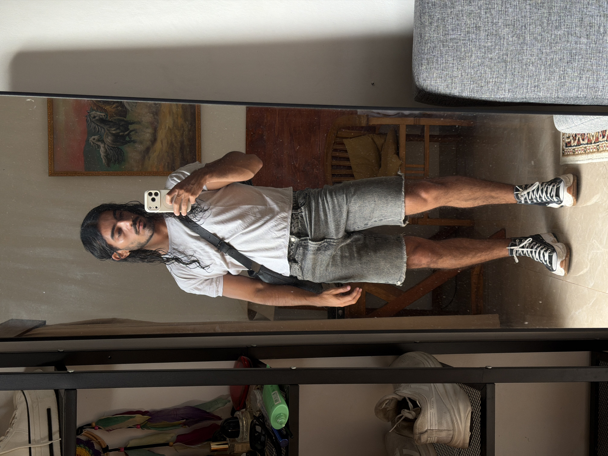 A man with long dark hair is taking a mirror selfie. He is wearing a white t-shirt, grey denim shorts, and black and white sneakers. He has a black crossbody bag slung over his shoulder. The mirror reflects a room with a painting on the wall, a wooden table, and a grey couch. Below the mirror is a black shelving unit with various items, including shoes, toiletries, and colorful cloths. The location is implied to be North Kuta, Indonesia.