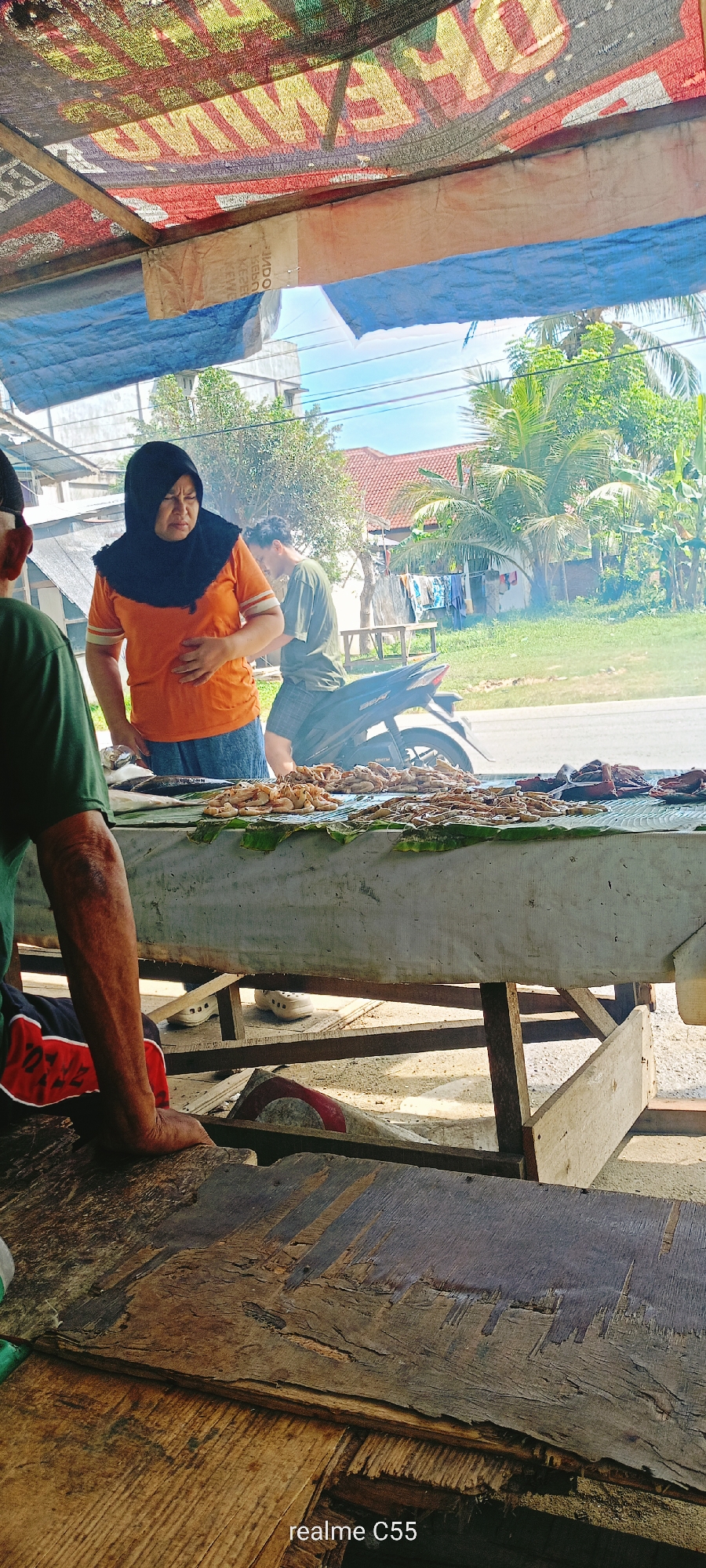 This media file captures an outdoor market scene in Lhokseumawe, Indonesia, characterized by a bustling street food stall under bright daylight. The central focus is a woman wearing a black hijab and an orange t-shirt, standing behind a makeshift wooden stall. Her left hand rests on her stomach, and she appears to be looking down at the items displayed on the table, with a somewhat pensive or absorbed expression.
The stall itself features a white tarp cover and displays a variety of fresh seafood, including numerous piles of shrimp, small fish, and what looks like grilled or dried fish, all arranged on banana leaves. In the mid-ground, a man in a green t-shirt and dark shorts is seen next to a black Honda Beat motorcycle, partially obscured by the woman. Another man, visible from the back in the left foreground, also wearing a green t-shirt, leans on a rough wooden surface.
The setting is an open-air environment with abundant tropical vegetation, including prominent palm trees, under a bright blue sky. Modest residential buildings are visible in the background, along with a paved road. Overhead, a makeshift canopy crafted from tarps provides shade; one tarp is blue, and another is a black mesh material with fragmented, inverted text, possibly from an advertisement, which includes partially visible words like "MIE," "INDOMI," "GEKBO," and "AWAS." Smoke rising in the background suggests active grilling or cooking. The overall impression is that of a lively, sunny day at a traditional market or roadside eatery. A "realme C55" watermark is present in the bottom right corner of the image.