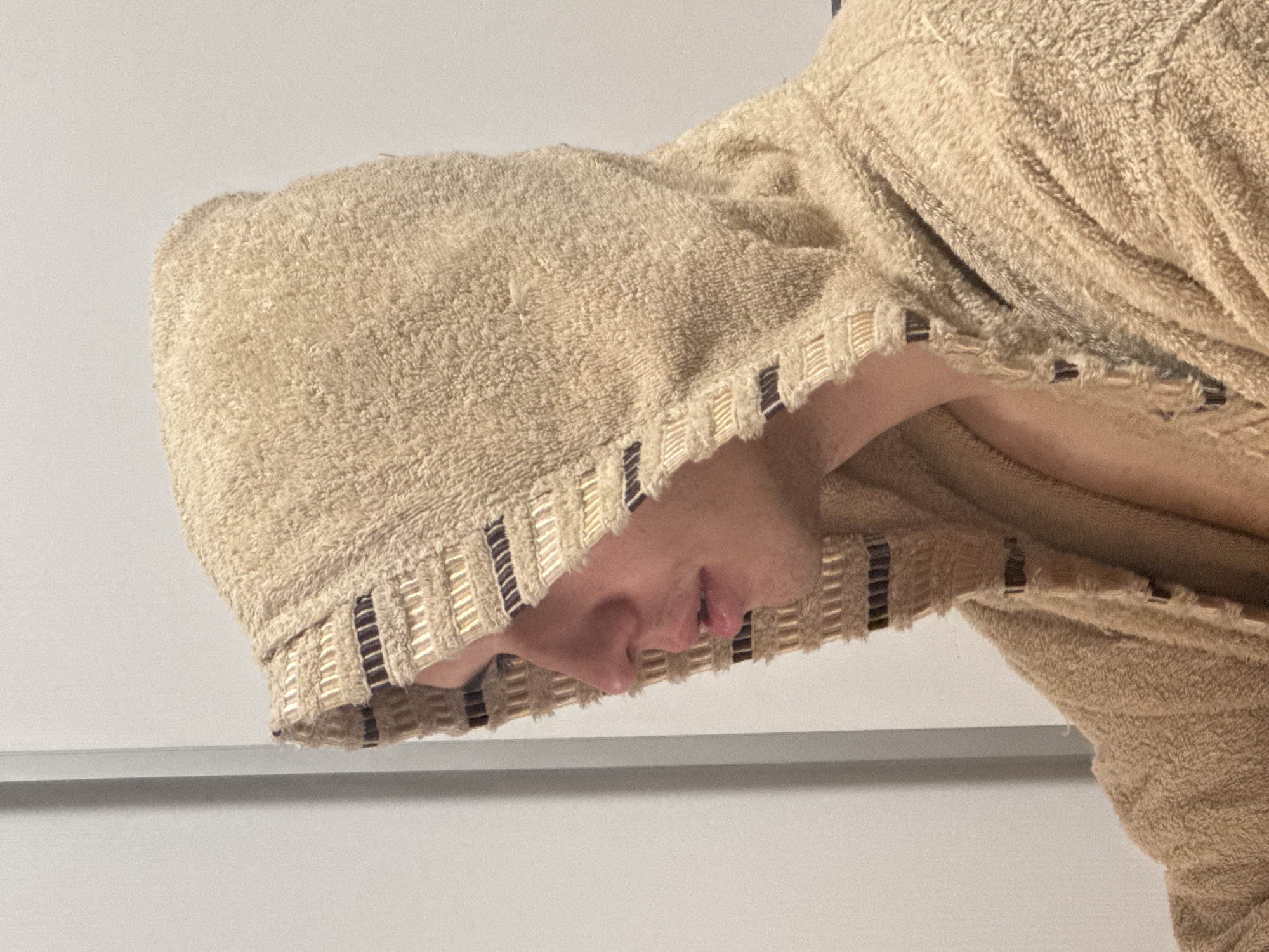 A male individual is depicted in a profile view from the mid-chest up, facing left. He is wearing a hooded garment, likely a bathrobe, made of a beige terry cloth material. The hood is drawn over his head, obscuring the top and back of his hair. The edges of the hood and the garment's front opening feature a decorative woven trim with alternating horizontal stripes of light gold and dark brown. His skin tone is fair, and his lips are slightly parted. The background consists of a plain, light-colored wall, with a subtle vertical seam or edge visible on the left side. The lighting is soft and even. The exact location, including the city of Sofia, Bulgaria, cannot be determined from the visual evidence provided in the image.