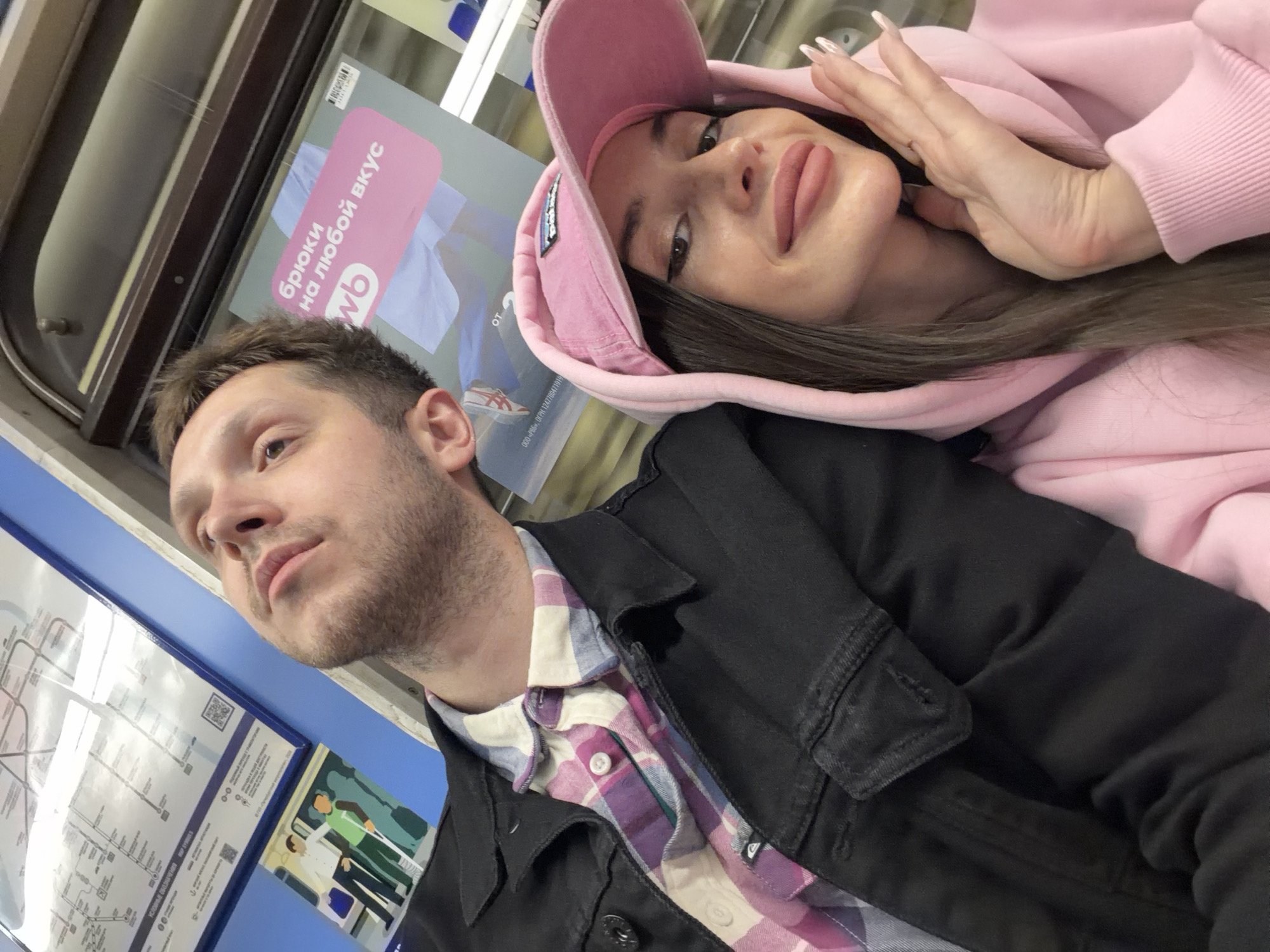 The image captures a man and a woman in what appears to be a subway car, likely in Saint Petersburg, Russia, given the presence of the Saint Petersburg Metro map on the wall. The man, with short brown hair and stubble, is wearing a plaid shirt under a black jacket. The woman, wearing a pink hooded sweatshirt and a pink baseball cap, has long brown hair. She is holding her hand near her face. An advertisement featuring the Russian e-commerce company Wildberries is visible in the background.