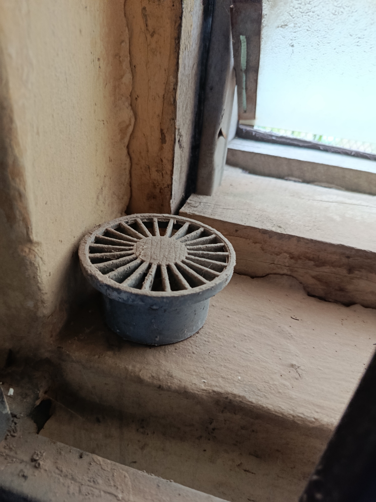 A dusty, circular drain cover with a radial pattern of spokes is positioned on a textured, sandy-colored windowsill. The drain cover is made of a grey, possibly metal or plastic, material and appears worn. Behind it, a wooden window frame shows signs of age and wear, with peeling paint or varnish. To the right of the drain, the window itself is visible, with frosted or textured glass obscuring the view outside, allowing only a diffused light to pass through. The overall environment suggests an older, possibly neglected building. This scene is located in Ukpor, Nigeria.