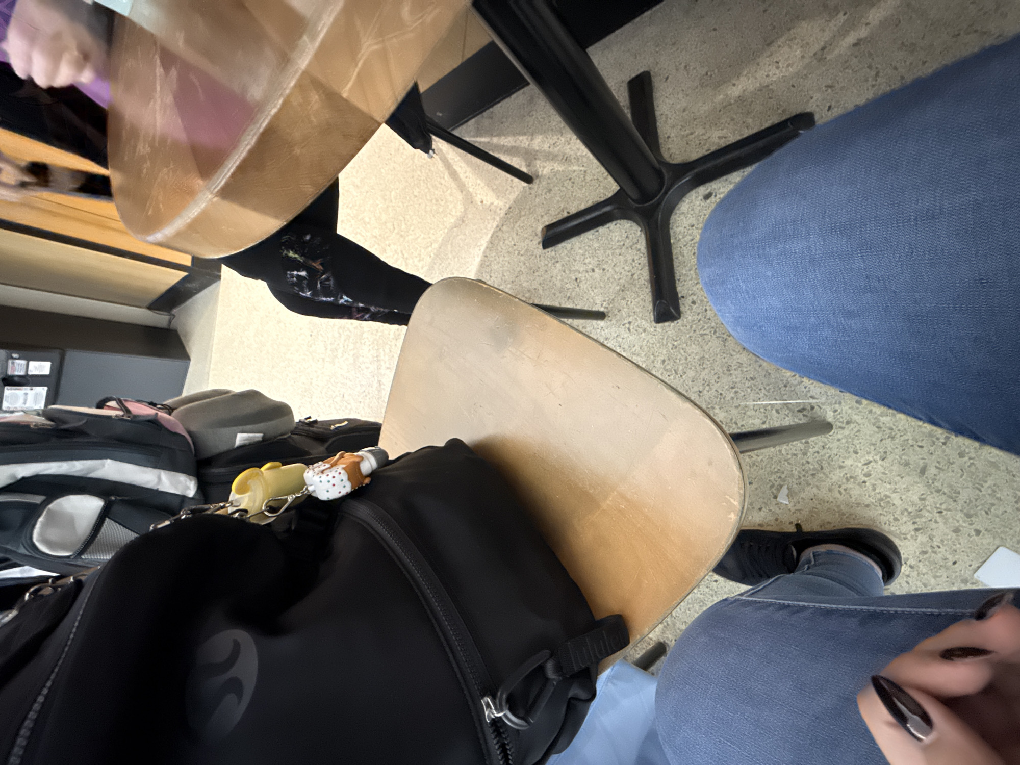 The image shows an indoor scene, likely a classroom or cafeteria, featuring two tables with people seated. One person is wearing black patterned leggings and another is wearing blue jeans. A black backpack with a keychain is sitting on one of the chairs. The environment consists of tiled flooring, with additional bags and items under one of the tables. Without additional information, determining a specific location in San Antonio, United States of America, is not possible.