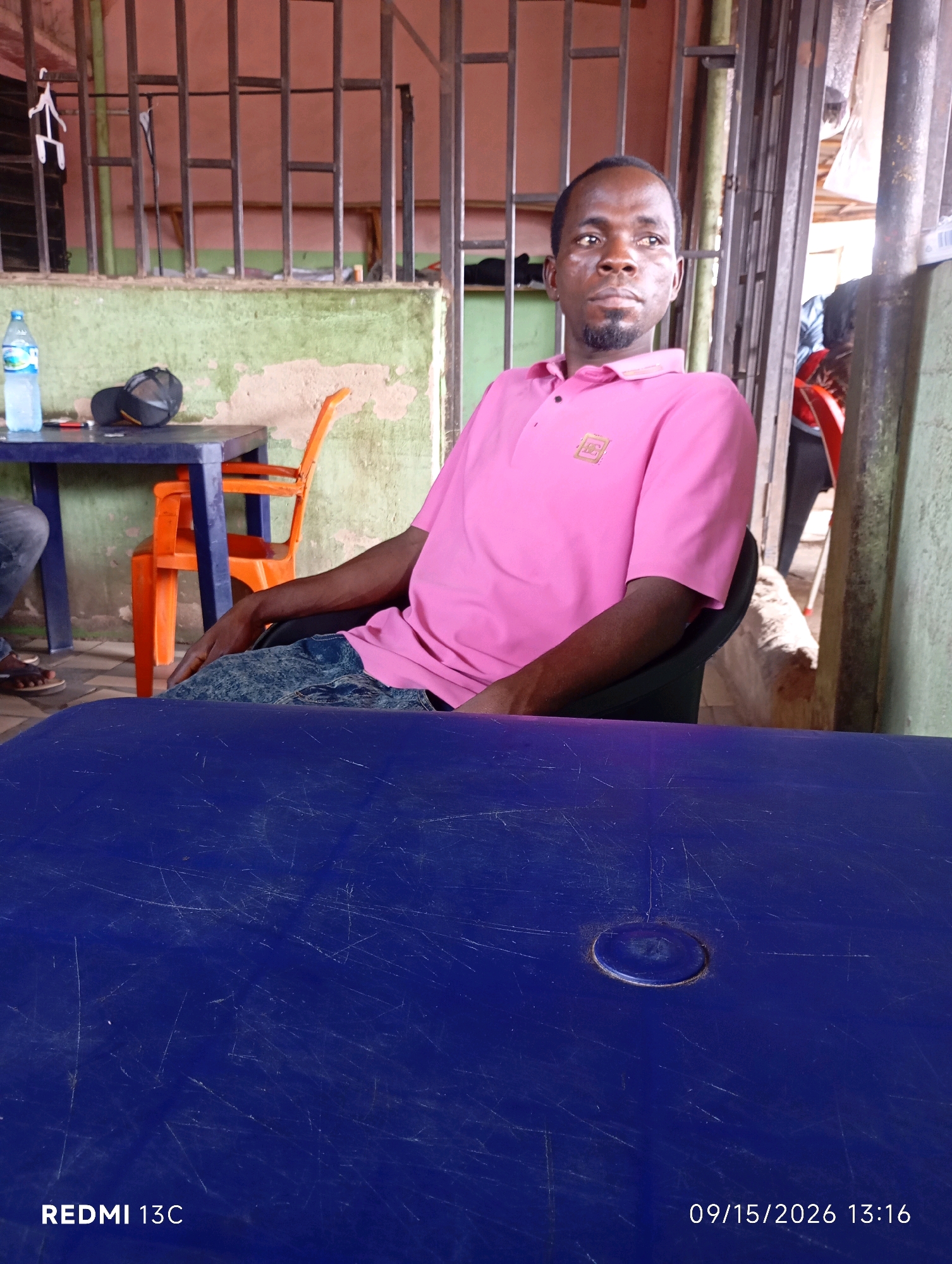The image depicts a man sitting at a table in a casual outdoor setting, possibly a food stall or eatery, in Shendam, Nigeria. The man is wearing a pink polo shirt and jeans. He is looking to his right with a neutral expression. In front of him is a blue table with some scratches. To his left, there is another table with a bottle of water and a cap on it. An orange plastic chair is visible behind him. The background shows a metallic structure and a green wall. The scene is illuminated by natural light. The overall impression is that of a candid moment captured in a local urban environment.