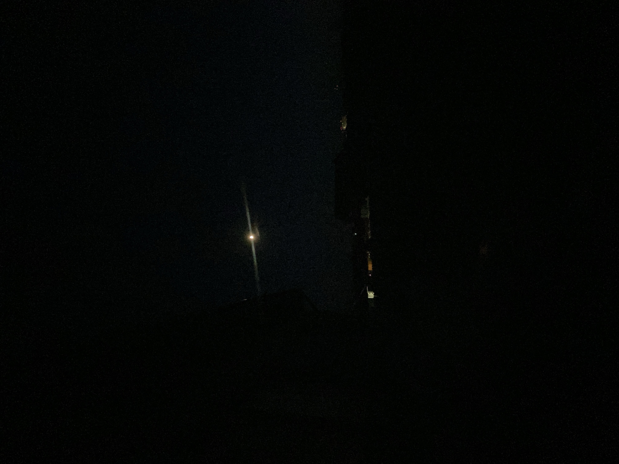 The image shows a dark, nighttime scene. A faint light source illuminates a pole-like structure in the distance. To the right, a building is partially visible, with some lights showing through the windows. The location is suggested to be Прикамский, Russia, based on provided metadata.