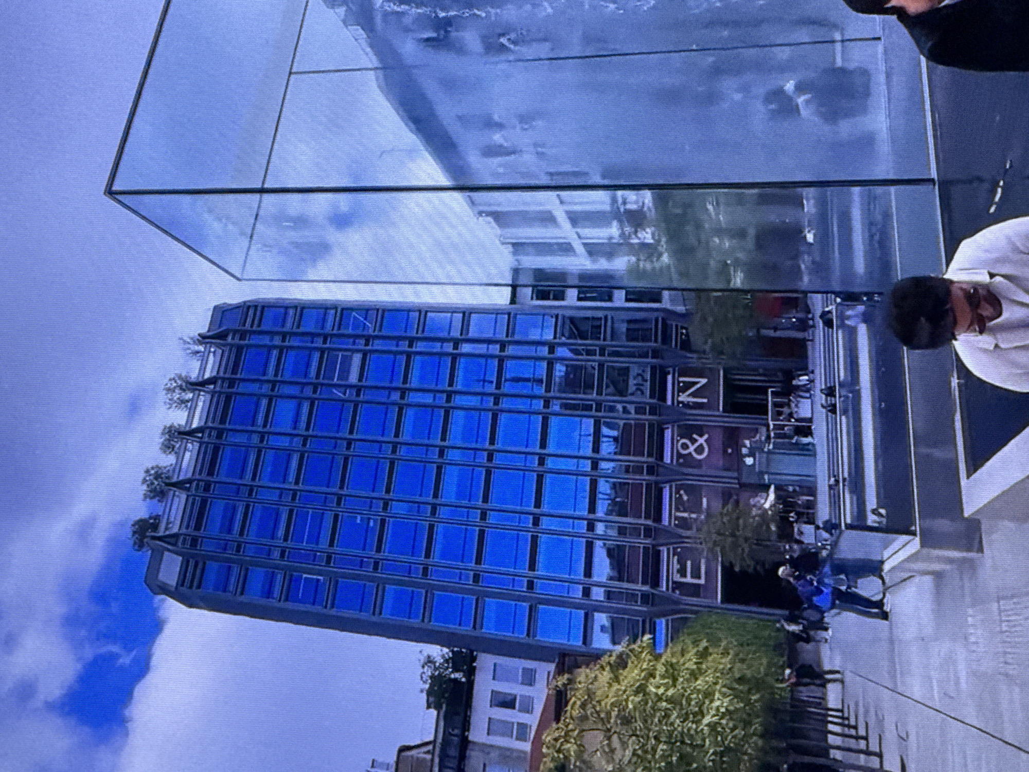 The image shows an outdoor urban scene with a modern blue glass building as the prominent subject. The building features a grid of windows and a distinctive tiered roofline with greenery. To the right, a large, transparent glass structure reflects the surrounding environment, including the blue building and the sky. In front of the buildings, there are several people walking and sitting, and trees are planted in rows along a paved walkway. The ground area appears to be a plaza with water features. The overall impression is one of contemporary architecture and public space within a city. The question asks for the location to be Dubai, United Arab Emirates, but the provided image does not contain any specific visual cues or text that would definitively place it in Dubai.