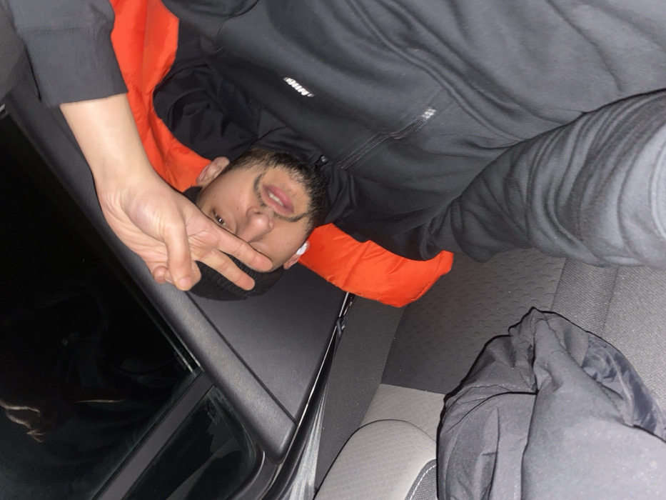 A person lies on their back, partially covered with an orange sleeping bag, in what appears to be the back seat of a car. They wear a black hooded sweatshirt, the hood up, and a black beanie. They are making a peace sign with their right hand, holding it in front of their face. Part of the car window and the rear seats are visible. The overall lighting suggests an indoor or nighttime setting.