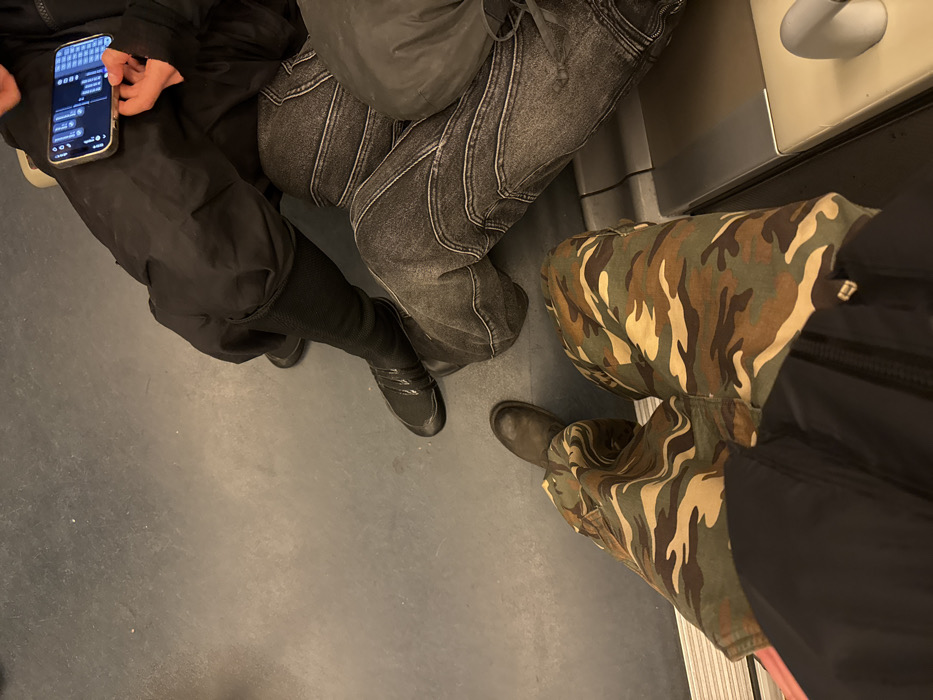 The image provides an overhead perspective of the lower bodies of three individuals seated within an RER train in Paris, France. The train interior features a uniform grey floor and beige bench seating with a light-colored metallic or plastic trim visible on the right.
In the immediate foreground, an individual, presumably the camera operator, is partially visible wearing camouflage-patterned cargo pants and one dark brown boot. Adjacent to them, another individual occupies the middle ground, wearing dark grey distressed denim jeans characterized by prominent horizontal seam detailing across the thighs and knees. A dark, possibly padded, jacket or bag is visible on this person's lap. Further back and to the right, a third individual is seated, wearing loose black trousers and dark, flat footwear. This person holds a smartphone horizontally with both hands, actively interacting with its display, which shows a text interface with a virtual keyboard. The smartphone is encased in a light-colored protective cover.