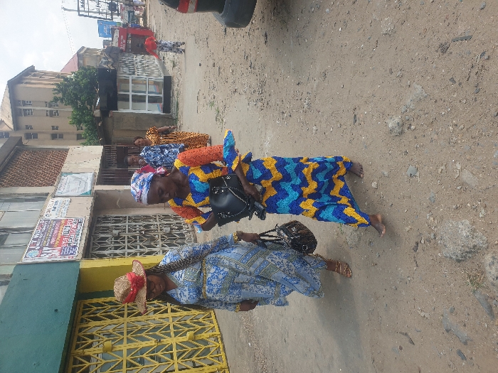 Several women stand on a dusty road. The woman in the foreground wears a light blue patterned dress, a straw hat, and carries a purse. Behind her, a woman in a blue and yellow patterned dress also carries a purse. Additional women in patterned dresses stand farther back near a building with barred windows and posters. A vehicle is partially visible at the top of the frame, with other buildings and power lines in the background.