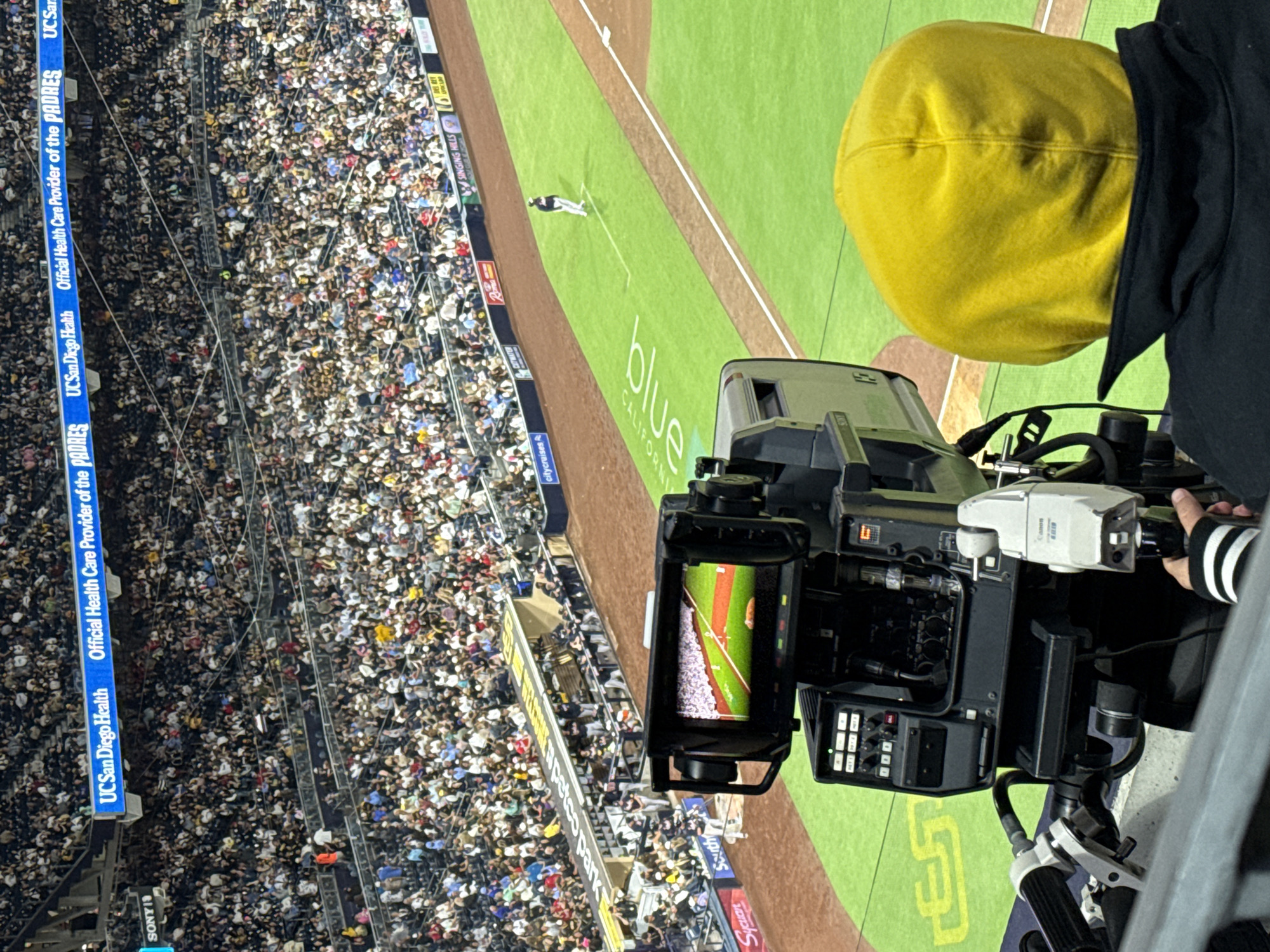 The image shows a baseball game at Petco Park in San Diego, California. A camera operator, wearing a yellow hoodie, is filming the game. The field is green with baseball markings, and a single player is standing on the grass. The stands are filled with spectators. In the background, banners with sponsors' logos are visible, including "UCSanDiego Health" and "Official Health Care Provider of the PADRES". The overall scene depicts a professional baseball event in a stadium environment.