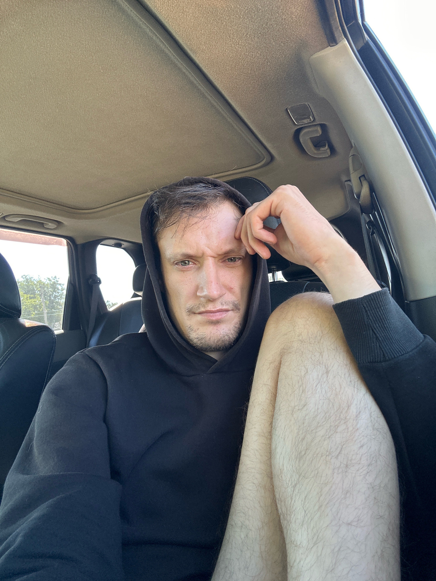 The image depicts a man in the driver's seat of a vehicle, with his head resting on his hand. He is wearing a black hoodie. The car interior is visible, including the headliner and a portion of the sunroof. The background shows trees and the sky through the car window, suggesting an outdoor location. The man's expression appears thoughtful or weary. There is no indication of the city of Makiv, Ukraine, or any other specific location.