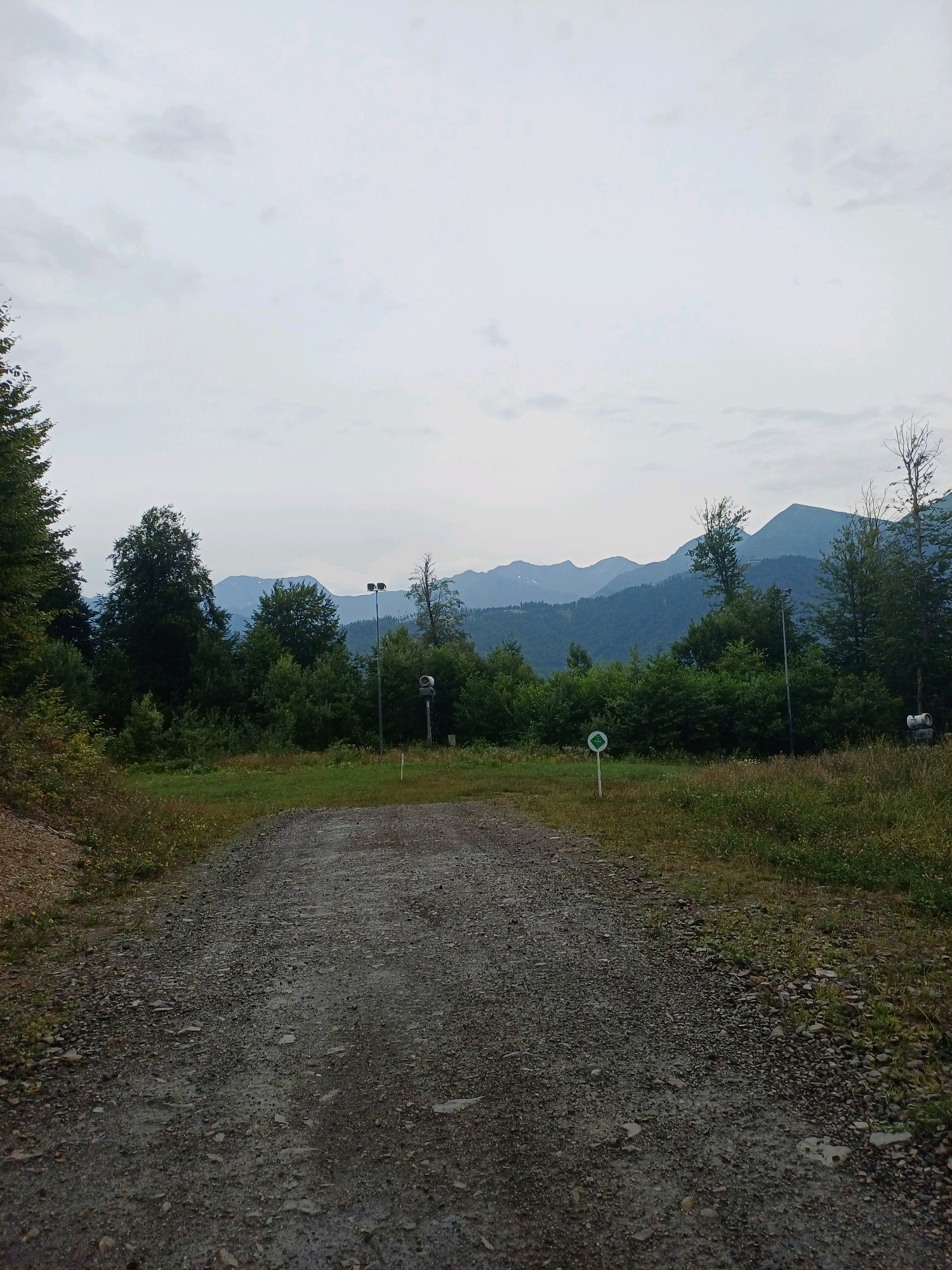 The image depicts a gravel path leading through a landscape of trees and mountains, likely in Estosadok, Russia. The path is wide and appears to be unpaved, with loose stones and dirt. On the left side of the path, there is a grassy slope with some dry vegetation. On the right side, there is more lush green grass and scattered wildflowers.
In the background, a dense forest of various trees stretches across the mid-ground. Beyond the trees, a series of majestic mountains rise, their peaks shrouded in a soft, hazy atmosphere. The sky above is overcast with a pale, diffused light, suggesting a cloudy day.
Several man-made objects are visible in the scene. On the right side, near the edge of the forest, a tall pole with two spotlights is erected. Next to it, there is a sign with a white circle and a green symbol inside, possibly indicating a trail or a route. Further to the right, another structure, partially obscured by trees, appears to be a piece of equipment or infrastructure. A white marker post is also visible in the grass on the right.
The overall impression is one of a natural, somewhat wild environment, likely a tourist or recreational area in a mountainous region. There are no people visible in the image, and no animals. The scene is static and peaceful, with a focus on the interplay between the man-made elements and the natural landscape.