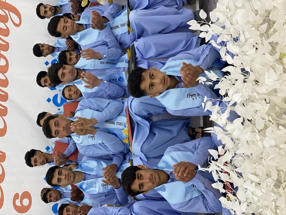 The image displays approximately twenty young men and male adolescents seated in rows at what appears to be a formal ceremony for Risha Professional Studies in Kandahar, Afghanistan. Each individual wears a light blue shalwar kameez and a light blue satin sash inscribed with "RISHA PROFESSIONAL STUDIES" and "ریشه" (Risha in Pashto/Dari). Most participants are captured with their hands raised together, palms open upwards, in a prayer or supplication gesture. Red folders or diplomas are visible on the laps of some individuals. A large white banner with red lettering, including "16" and parts of "CEREMONY", forms the background. The foreground features a decorative arrangement of white, artificial foliage. The individuals' expressions are generally serious or contemplative.