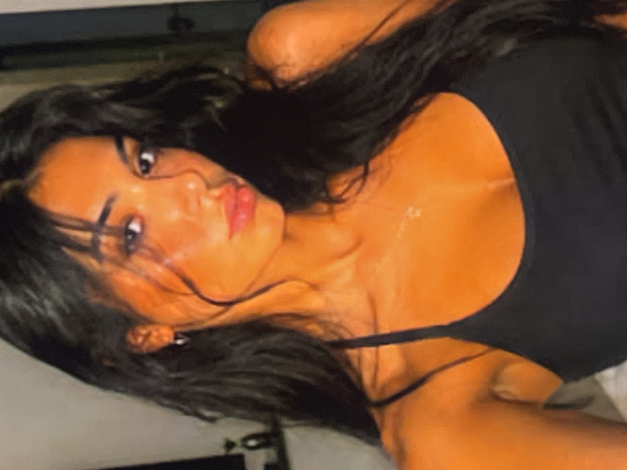 A close-up, slightly blurry, selfie of a young woman with long, dark, wavy hair. She is wearing a black tank top and a delicate necklace. The lighting is warm and orange-toned, giving her skin a tanned appearance. Her hair falls around her shoulders and frames her face. The background is dark and out of focus, with a hint of a shelf and an object on it visible on the left side. The woman is looking directly at the camera with her lips slightly parted.