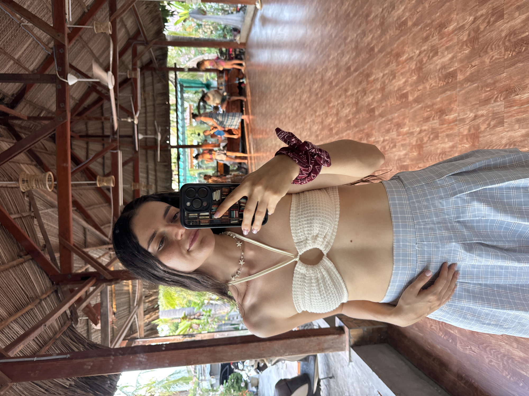A young woman with long dark hair takes a mirror selfie in what appears to be a yoga studio in Mũi Né, Vietnam. She is wearing a white crocheted halter top and a light blue plaid skirt. She holds a black smartphone, decorated with images of books, in her right hand. A maroon scrunchie adorns her wrist. The environment features a traditional Vietnamese structure with a thatched roof, wooden beams, and a polished wooden floor. In the background, other individuals engage in yoga activities within the open-air studio. Lush greenery is visible outside the studio. Ceiling fans are suspended from the roof structure.