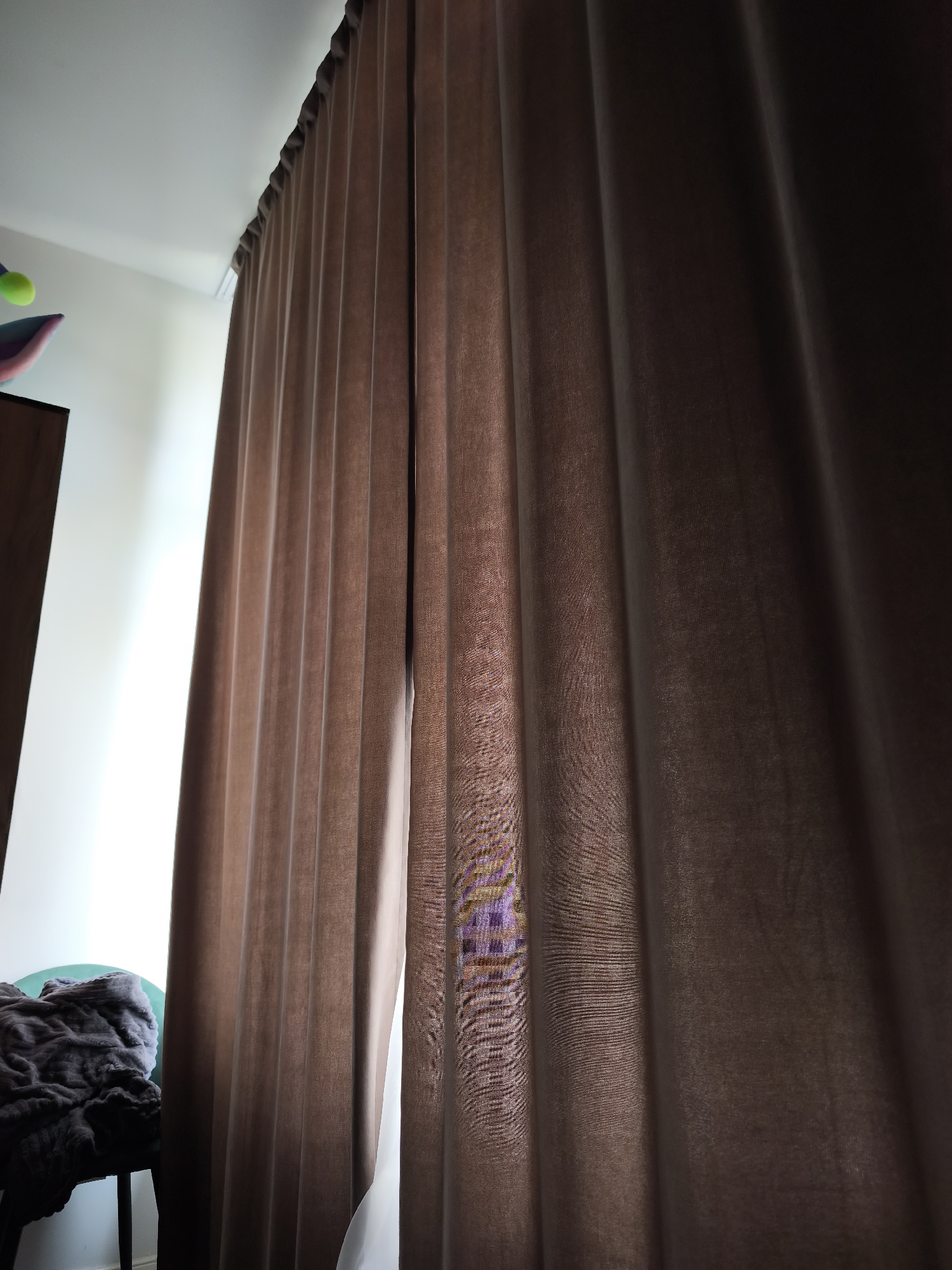 A close-up, indoor shot depicts heavy, dark brown curtains hanging from a ceiling track, with a slight gap revealing a glimpse of the window behind and some light. To the left of the curtain, a small portion of a room can be seen, including part of a green chair covered with a dark grey blanket. No people are visible in the image. There is no indication of Moscow, Russia.