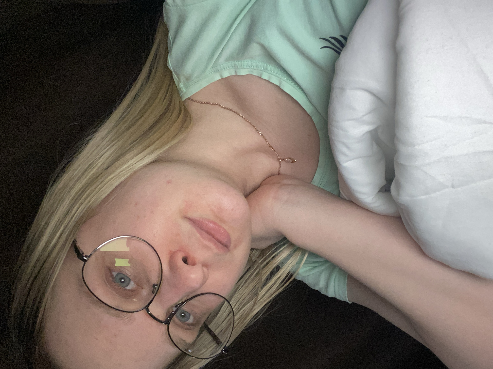 An individual is depicted in a close-up, dimly lit indoor setting in Chaikovsky, Russia. The subject is a female with fair skin, long blonde hair, and blue eyes, wearing round, dark-framed spectacles that reflect a rectangular light source. Observable skin imperfections, including redness, are present on her face. Her expression is neutral with slightly parted lips. She wears a light green short-sleeved shirt with an indistinct dark marking on the lower right, and a thin, rose gold chain necklace featuring a small teardrop pendant. The subject's head is tilted, resting on her right hand or forearm. The immediate environment includes a dark, textured background (likely upholstery) on the left and top, and white fabric (consistent with bedding or a pillow) on the right and bottom.