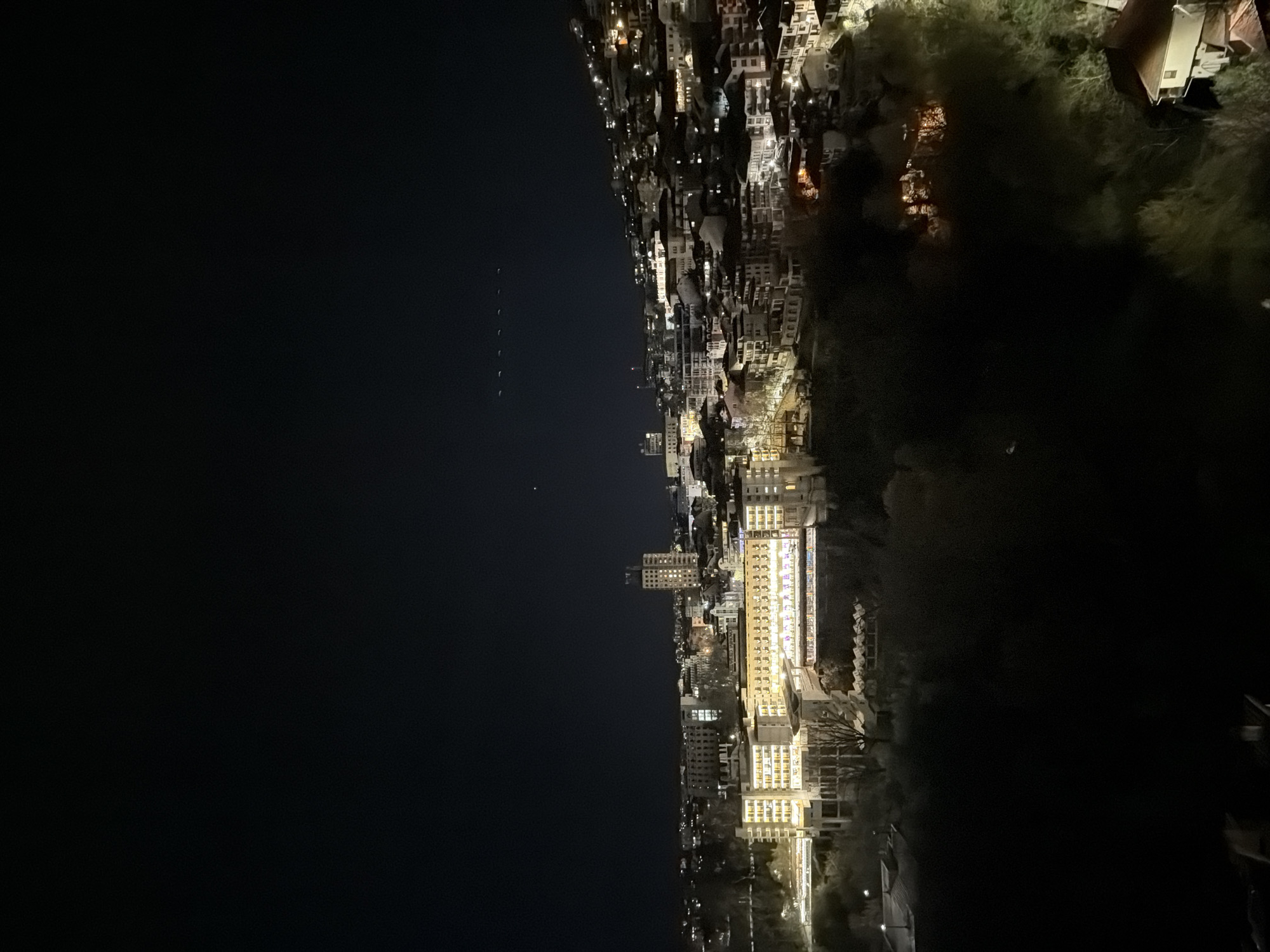The image shows a nighttime view of the city of Veliko Tarnovo, Bulgaria. On the right side of the frame is a cluster of buildings nestled into a hillside, their illuminated windows creating a network of light. A large building stands out, its facade brightly lit with rows of windows. Further back, the lights from other structures spread along the terrain, blending into the natural landscape. The left side of the image is dominated by the black sky and darkness.