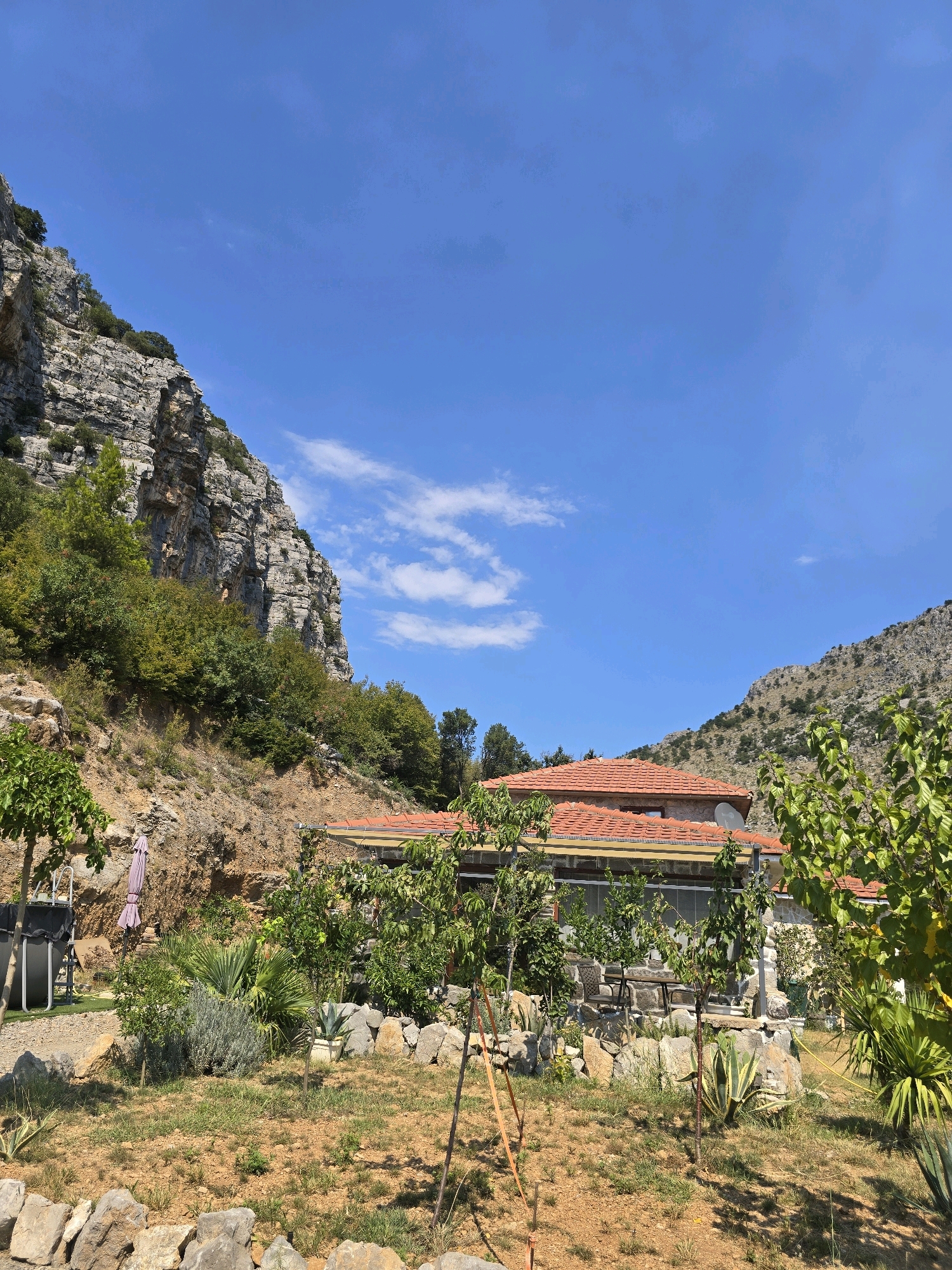 The image depicts a rural landscape with a house nestled against a rocky mountainside. The house has a red-tiled roof and stone walls, with a satellite dish visible on the roof. The surrounding environment is characterized by dry, rocky terrain with sparse vegetation, including trees, shrubs, and succulents. In the foreground, there is a garden area with a stone wall. A small above-ground pool and a patio with outdoor seating are also visible. The sky is clear blue with a few wispy clouds. The scene suggests a warm, sunny day in a mountainous region. The specific location within Podgorica, Montenegro, is not evident from the image itself but the style of the house and vegetation is consistent with the mountainous regions surrounding the city.