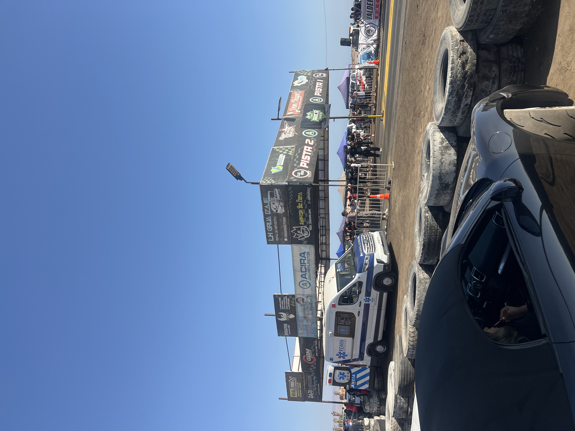 The image depicts a scene at a drag racing event in Arica, Chile. The sky is clear and blue. A dark car is prominently positioned in the foreground, with a person visible inside the driver's seat. Along the racetrack, there is a barrier of stacked tires. Beyond the barrier are promotional banners advertising businesses and events, including "PISTA 2," "ACIRA REPUESTOS," and "LH GRUA IZAJE." An ambulance is parked nearby. Spectators and event staff are visible behind a safety fence. The overall environment suggests a public gathering for a motor sports event.