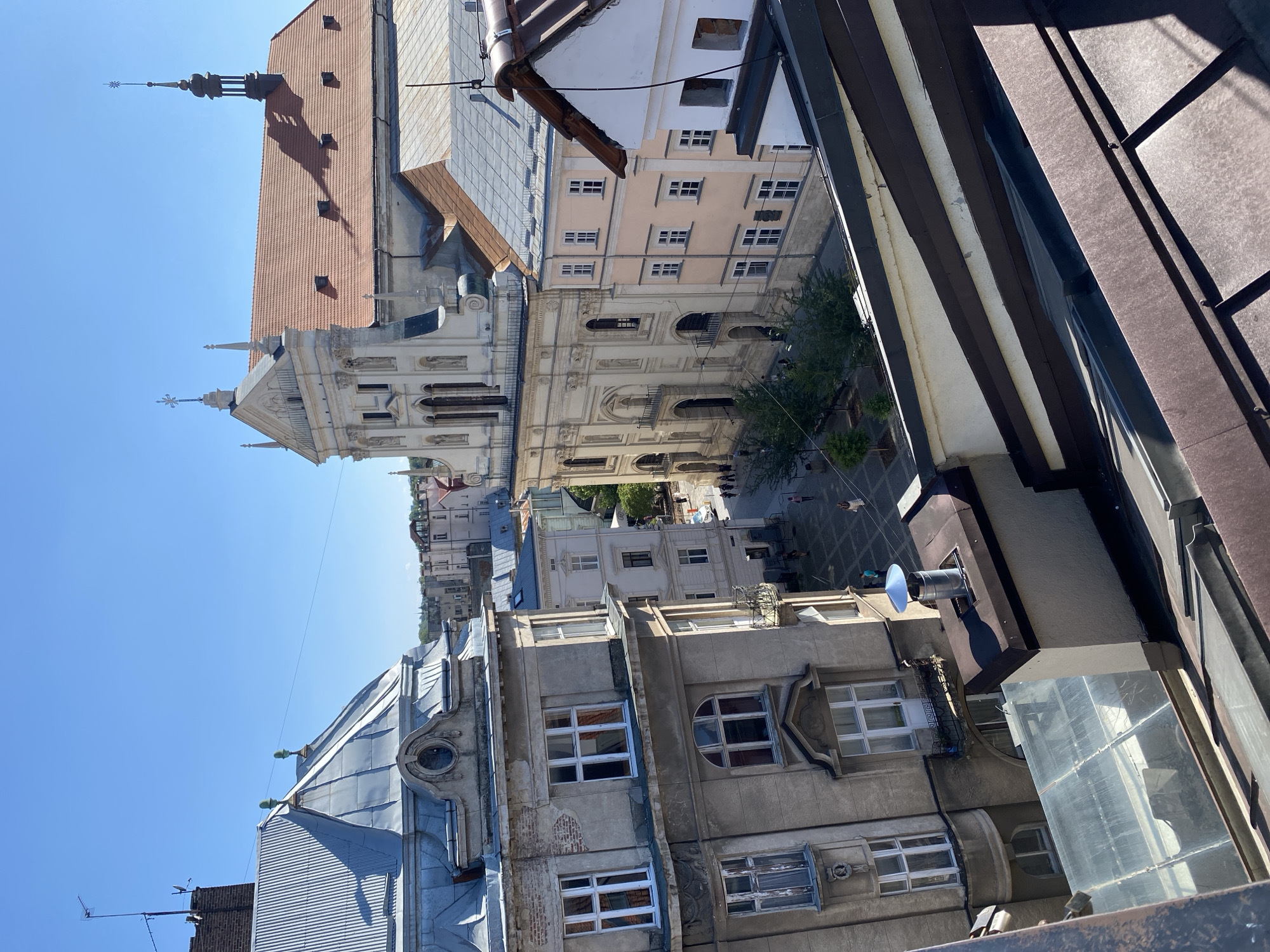 An aerial view captures a sunlit urban scene in Lviv, Ukraine. In the midground, a grand, ornate cream-colored building with a red-tiled roof and spires dominates the skyline. To its left, a grey, weathered building with a mansard roof and numerous windows stands prominently. In the foreground, the dark, sloped roofs of several buildings create a layered effect. A paved square with scattered trees and a few pedestrians is visible between the main buildings. The sky above is a clear, vibrant blue.