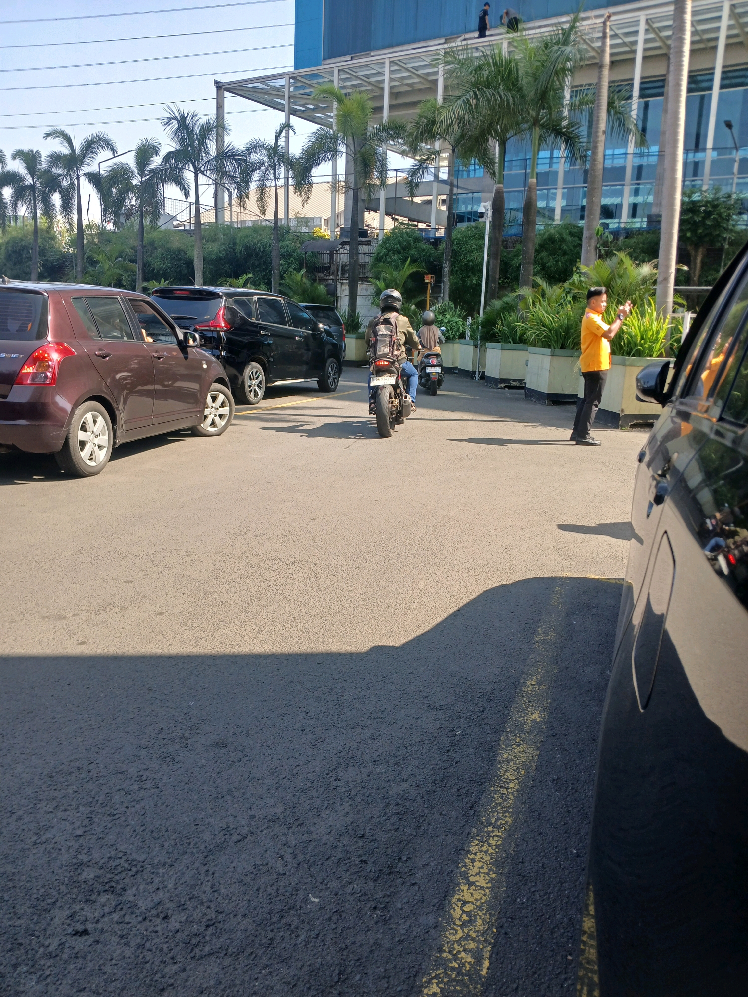 The image shows a parking lot with several vehicles and people. In the foreground, a dark-colored car is partially visible, with its side and rear passenger door in view. Further into the scene, a brown sedan is parked, with its front and side visible. Behind the brown sedan, a black SUV is parked. Two individuals on motorcycles are on the road. One motorcycle rider, wearing a brown jacket and a helmet, is in the center of the frame, moving away from the camera. Another person on a motorcycle is behind the first. A man in an orange shirt and black pants is standing on the right side of the road, looking at his phone. In the background, there is a modern building with blue glass windows and a white roof structure. Several palm trees and other greenery are scattered around the area. The scene appears to be outdoors during the daytime, with clear skies. The ground is paved asphalt. This scene is consistent with a parking area in an urban environment, possibly near an office building or commercial complex.