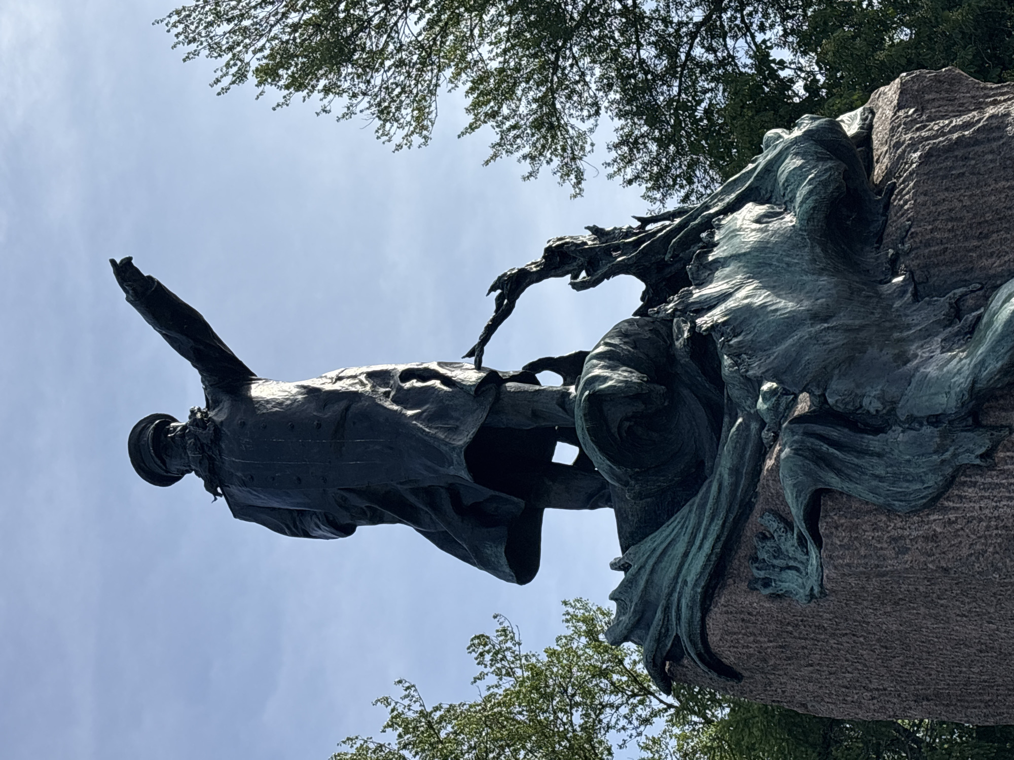 The image shows a bronze statue of a man in a long coat and a cap, pointing his right arm forward and upward. The statue stands on a stone pedestal that is adorned with abstract bronze sculptures resembling waves and perhaps sea creatures. The statue is dark in color and appears to be weathered. Green trees with dense foliage are visible on the right and left sides of the frame, against a clear blue sky with scattered clouds. The perspective is from a low angle, looking up at the statue. This is a memorial to Admiral Fyodor Ushakov, located in the city of Kronstadt, Russia.