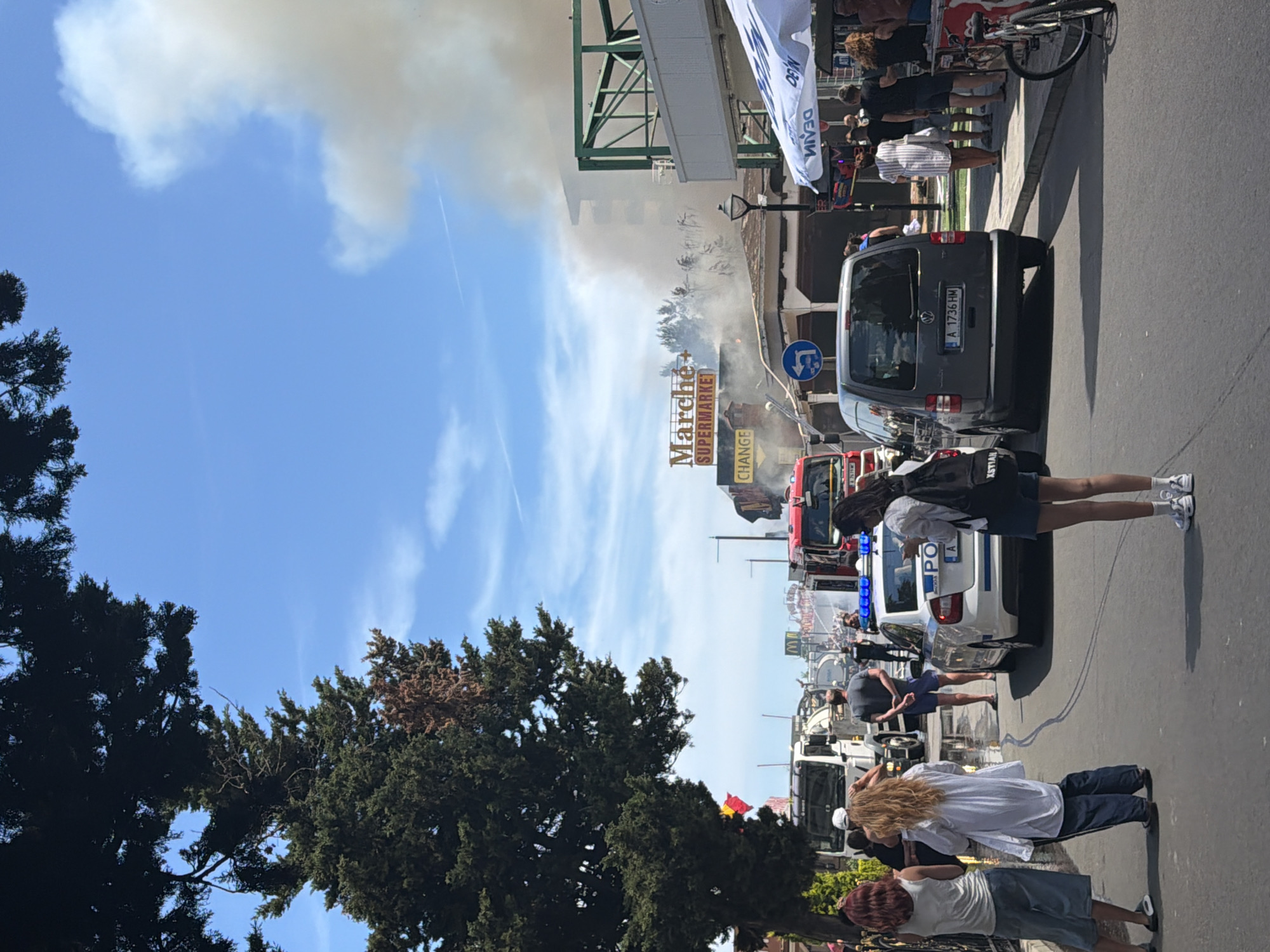 The image depicts a scene in the city of Nesebar, Bulgaria, featuring a significant fire. Thick smoke billows from a multi-story building on the right side of the frame, obscuring parts of the structure and casting a haze over the sky. A prominent sign reading "Marche SUPERMARKET" is visible on the building. In the foreground, a street is lined with emergency vehicles, including at least two fire trucks, one red and one white with a water tank, and a police car with its blue lights illuminated. Several individuals are present, some observing the fire, others appearing to be emergency personnel or bystanders. A group of people stands on the left side of the street, some looking towards the incident. Further down the street, more vehicles and people are visible. The environment suggests a sunny day with a clear blue sky, contrasted by the dark smoke from the fire.