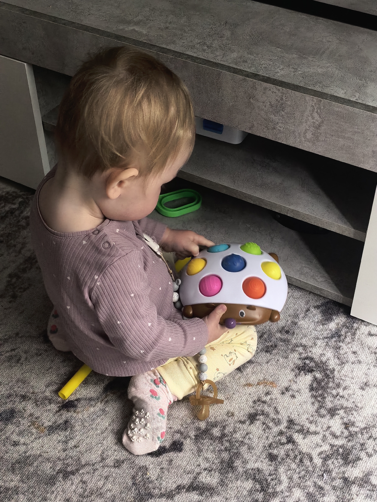A young child with light brown hair is seated on a grey and white patterned carpet, engrossed in playing with a colorful toy. The child is viewed from behind and slightly to the side, wearing a ribbed, long-sleeved purple top adorned with tiny white heart patterns, yellow pants featuring animal prints, and light pink socks with anti-slip white grips and small floral designs. A beaded pacifier chain with a wooden pacifier hangs from the child's neck/clothing.
The toy the child is holding is a brown and white interactive object, possibly a hedgehog or turtle, featuring multiple colorful, round buttons or balls in yellow, blue, pink, orange, and green on its back. The child's hands are actively engaging with these colorful elements.
Behind the child is a low, multi-tiered shelving unit or TV stand with a dark grey, textured, concrete-like finish. Underneath the bottom shelf, a bright green ring-shaped object and a small white container with a blue lid are visible. A yellow plastic tube-like object lies on the carpet near the child's left leg. The carpet itself shows some brownish stains. The setting appears to be indoors, likely a home environment, in Sterlitamak, Russia. No specific time of day, weather, or visible text can be determined from the image.