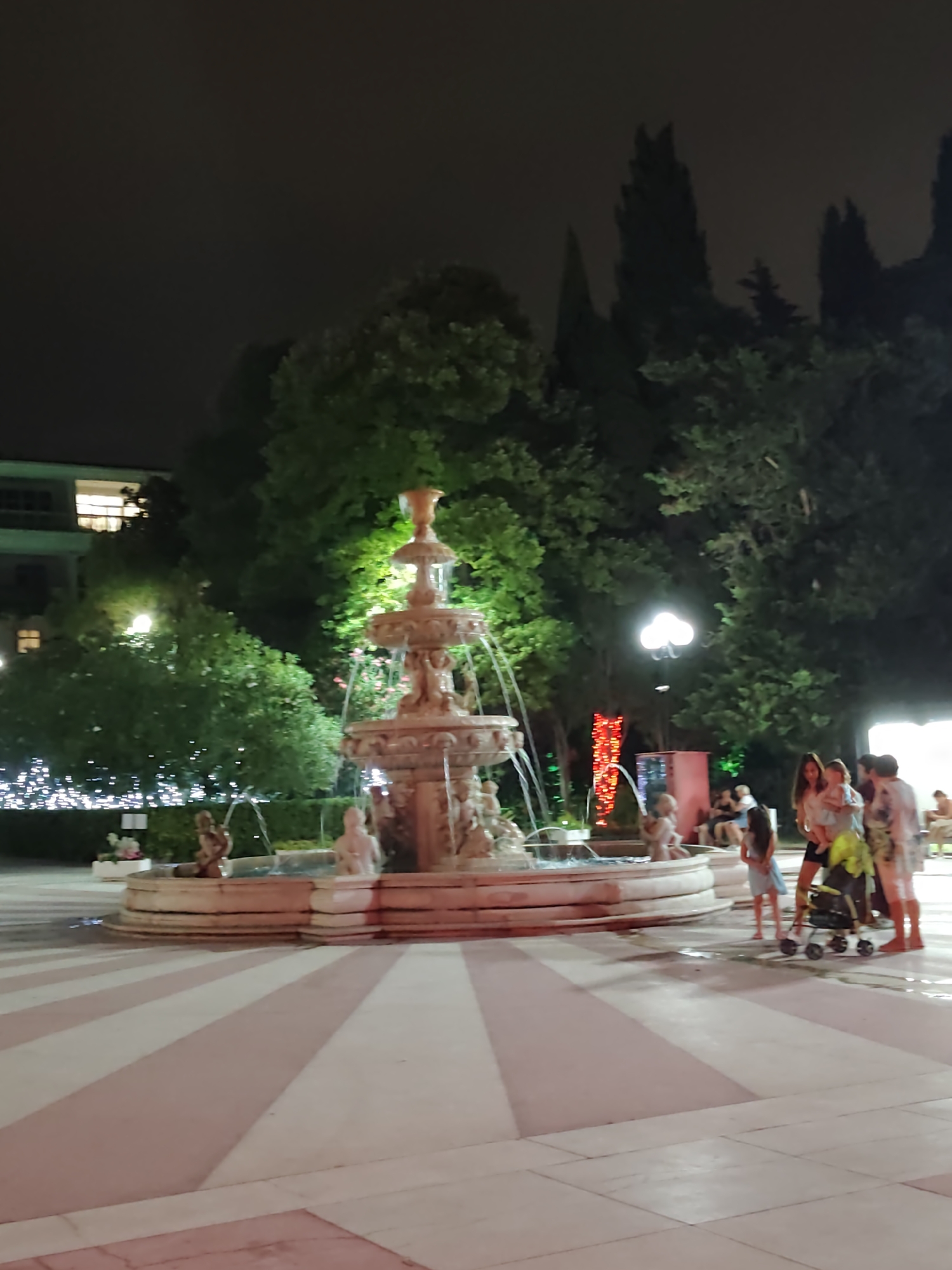 A night scene in Sochi, Russia, shows a fountain in a plaza. The fountain, made of a light-colored stone, has multiple tiers and is illuminated by artificial lights, causing water to spray from it. Around the fountain, there are several people, including a group of women and children, some of whom are standing near a stroller. The plaza floor has a distinctive pattern of pink and white stripes radiating outwards from the fountain. In the background, there are trees, some decorated with string lights, and a building with illuminated windows. The overall atmosphere is festive and illuminated.