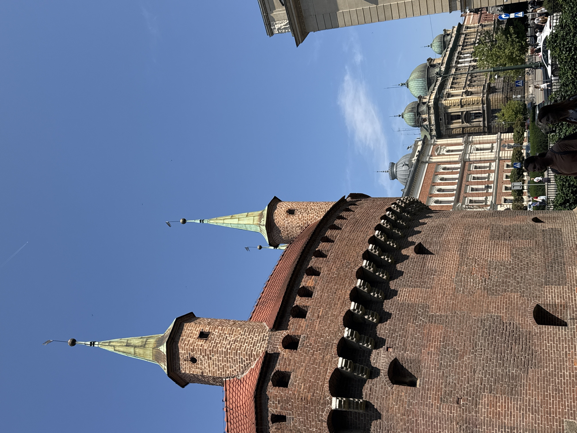 The image shows a low-angle view of the Krakow Barbican, a historic medieval fortification in Krakow, Poland, on a clear day. The barbican, constructed from weathered red brick, features a rounded wall with arched openings and crenellations along the top. Two conical green-roofed turrets rise from the structure. In the background, a row of ornate historic buildings with green domed roofs and classical architectural elements line a street. A white car is visible on the street, and several people are walking on the sidewalk. The sky is a vibrant blue with a few wispy clouds and a faint contrail from an airplane.