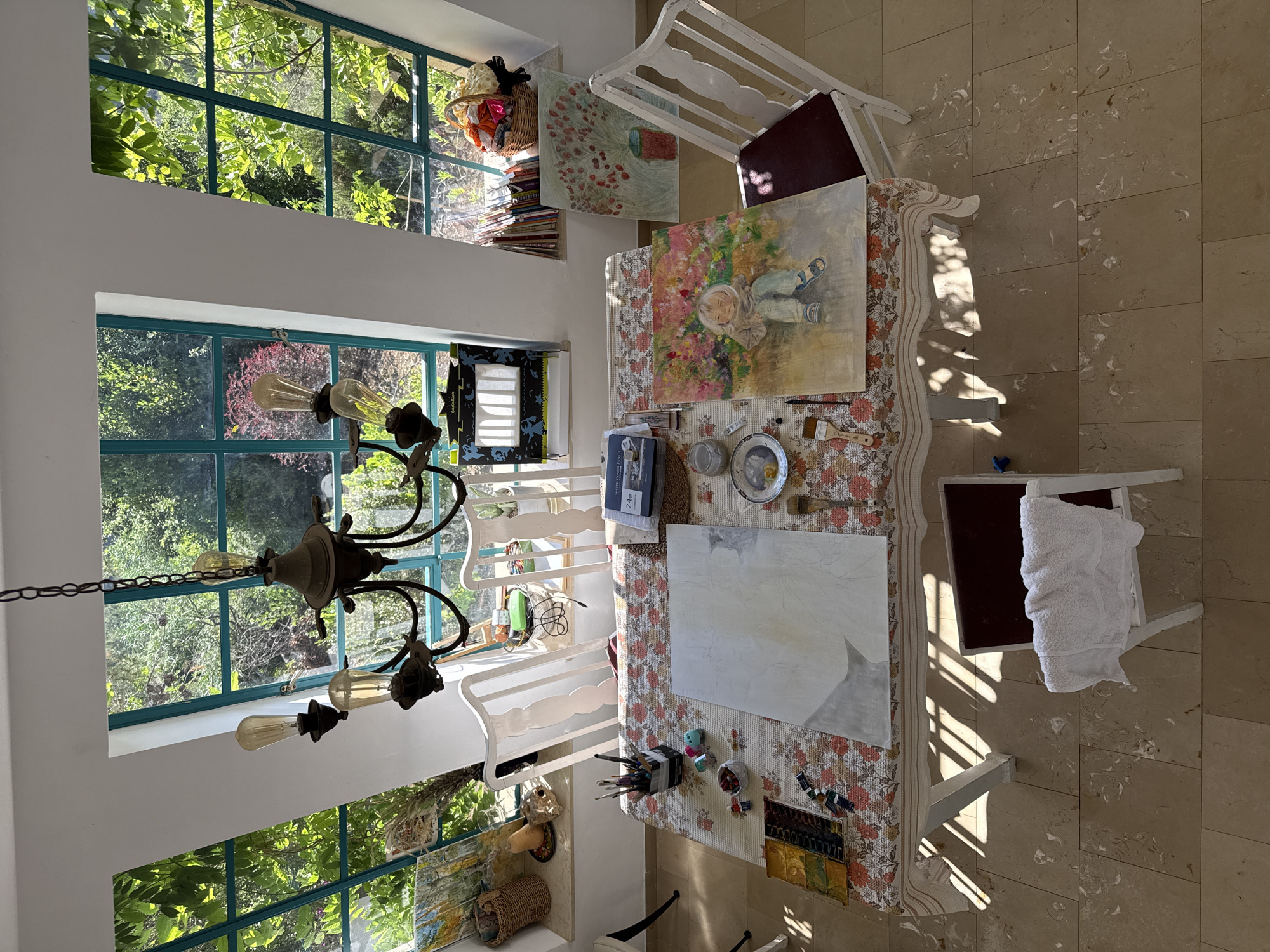The image depicts an art studio space, likely in Kfar Vradim, Israel, as suggested by the presence of large windows with a view of abundant green foliage, characteristic of the region's natural landscape. The scene is centered around a dining table that has been repurposed as an art workstation.
On the table, two canvases are prominently displayed. One is a large, unfinished abstract painting with subtle grey and white tones, sketched with faint lines. Next to it, a completed painting of a young child sitting amidst a vibrant floral background is propped up. Various art supplies are scattered across the table, including a palette of watercolors, paint tubes, brushes in a holder, a jar of what appears to be medium, and a small dish with paint. A book titled "WATER COLOR COLOR PENCIL" is also visible, along with a small plate holding what look like art tools and a tiny toy car.
The table is covered with a floral-patterned tablecloth. Several white wooden chairs are positioned around the table. One chair, to the right of the table, has a dark red cushion. Another chair, at the bottom center of the image, has a white towel draped over its back.
Behind the table, a series of large, multi-paned windows with turquoise frames offer a view of lush greenery and trees, suggesting the studio is in a well-landscaped or natural setting. Sunlight streams through the windows, casting distinct patterns of light and shadow on the table and floor.
On a windowsill behind the table, there are more art supplies, including what appears to be a collection of books, a woven basket filled with colorful items, and other decorative objects like a squash and a skull-like carving. Above the table, a rustic chandelier with three light bulbs hangs from the ceiling.
To the left of the windows, a dark framed shadow puppet theater setup is visible. The floor is tiled with large, light-colored stone tiles, which are partially covered by shadows from the table legs and the sunlight. The overall impression is that of a functional and creative art space, bathed in natural light.