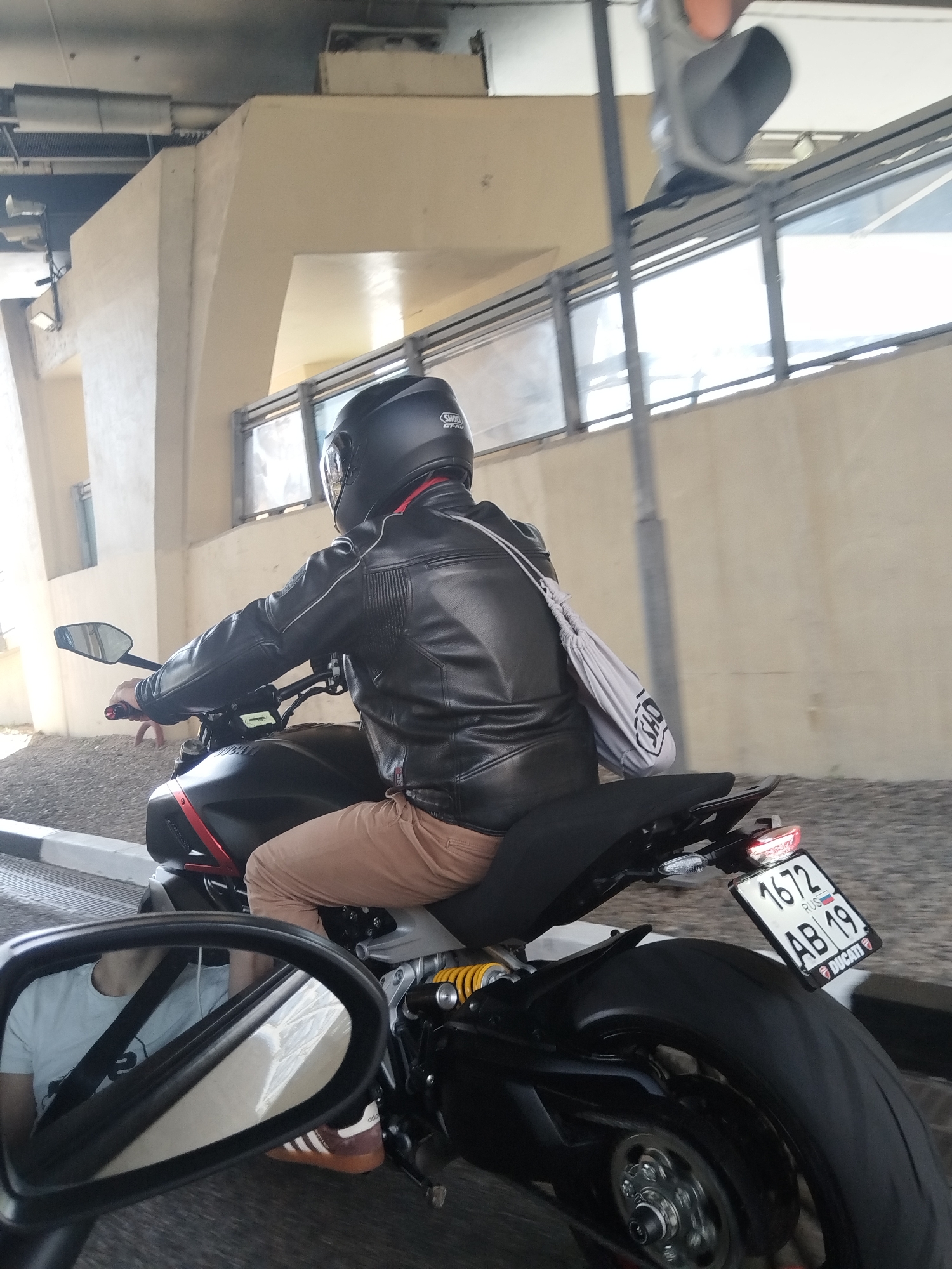 The image shows a person riding a black Ducati motorcycle on a road, as seen from the perspective of another vehicle's side mirror. The rider is wearing a black leather jacket, beige pants, a black helmet, and brown Adidas shoes. A gray shoulder bag is slung over their shoulder. The motorcycle has a license plate that reads "1672 AB 19 RUS", indicating it is registered in Russia. The setting appears to be in a city, evidenced by a large concrete structure with windows and railings in the background. The presence of a concrete support structure above suggests the scene is taking place under a bridge or an elevated road. The location is Sochi, Russia, as indicated by the Russian license plate. The environment is urban, with street-level infrastructure visible.
