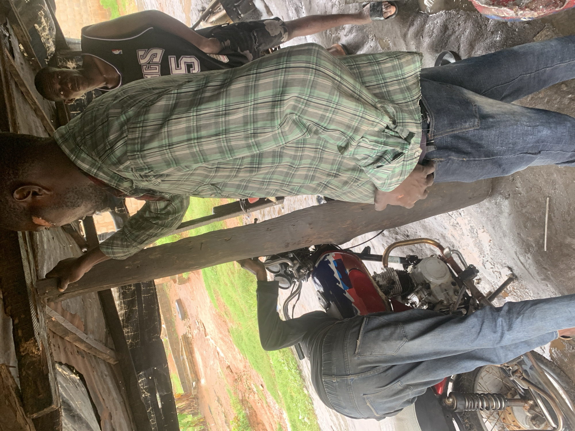 In Shagamu, Nigeria, two men are gathered under a makeshift shelter constructed from corrugated metal and rough wooden beams, a common sight in many parts of the city. One man, wearing a green and white plaid shirt and blue jeans, is bent over a red and blue motorcycle, his left arm extended as he appears to be working on the engine. The other man, dressed in a black basketball jersey with the number "5" and patterned shorts, stands observing him. The ground is wet and muddy, suggesting recent rain, and the surrounding area shows evidence of both natural elements and man-made structures, with glimpses of greenery and buildings in the background. The scene captures a moment of mechanical repair under less-than-ideal conditions, typical of informal workshops found within Nigerian cities.