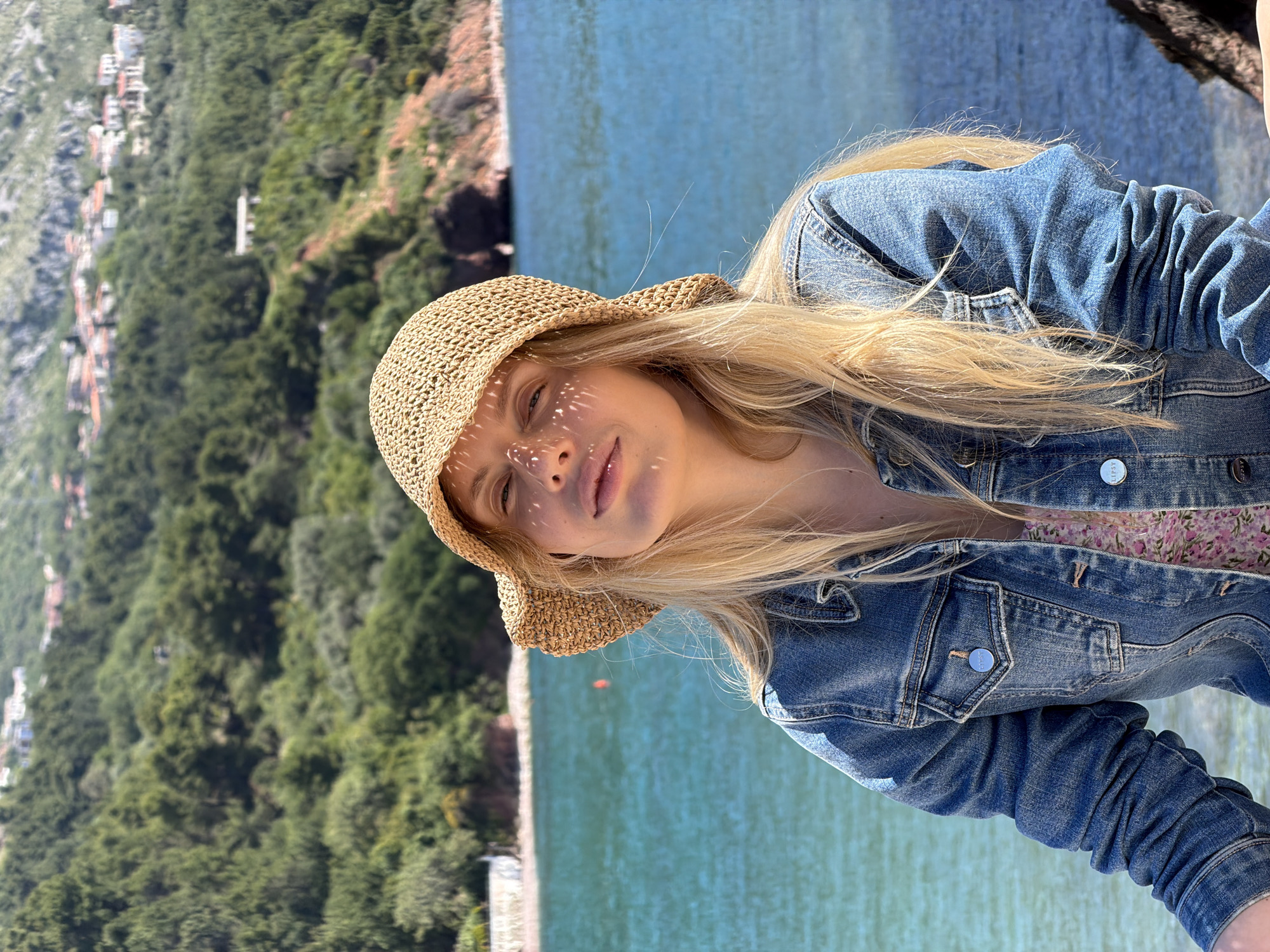 A blonde woman wearing a straw hat and denim jacket poses in front of the Adriatic Sea near Sveti Stefan, Montenegro. The scene includes a lush, green, mountainous landscape in the background, with glimpses of buildings and a beach along the coastline. The woman is the main subject, her face partially shadowed by the hat.