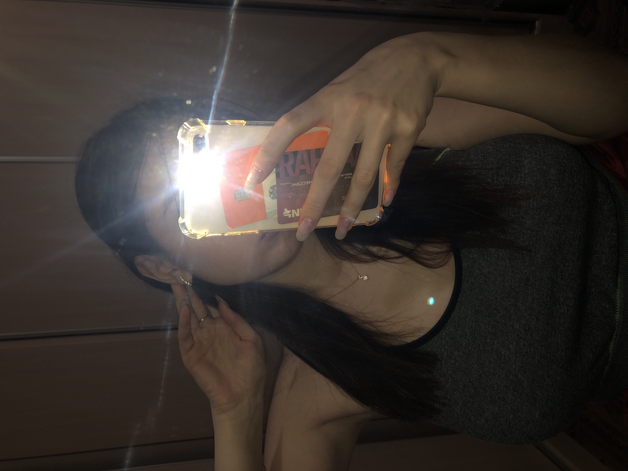 A woman is taking a selfie in a mirror. She is holding a phone with a clear case that contains cards, including one that appears to say "KAHRA". She is wearing a gray tank top. She has long dark hair, earrings, and a necklace. The reflection in the mirror also shows a light fixture. The location is unclear from the image and cannot be definitively determined to be in Noviy, Russia.